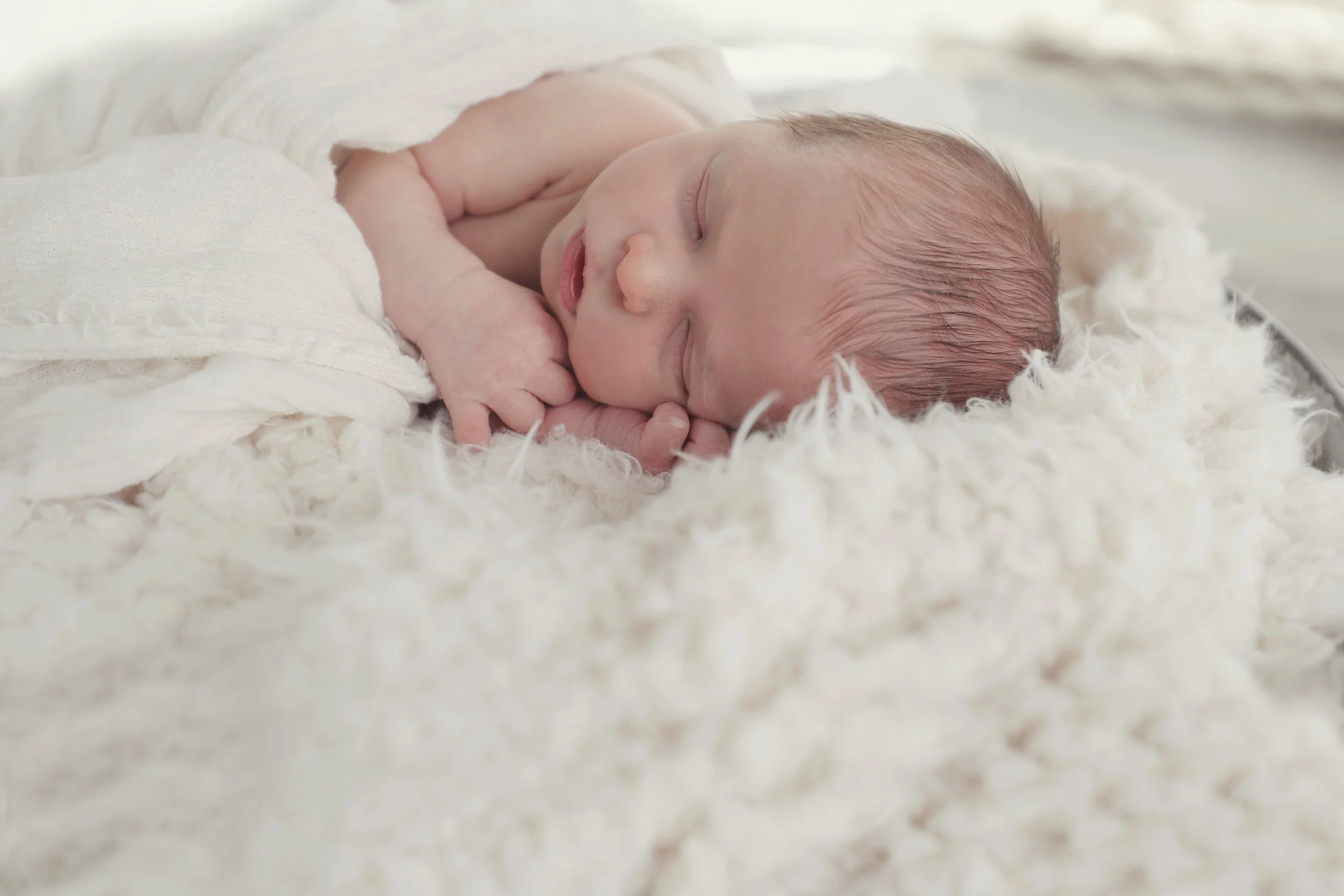 Close-up of a sleeping newborn baby lying on a soft, fluffy white blanket, wrapped in a light-colored cloth.