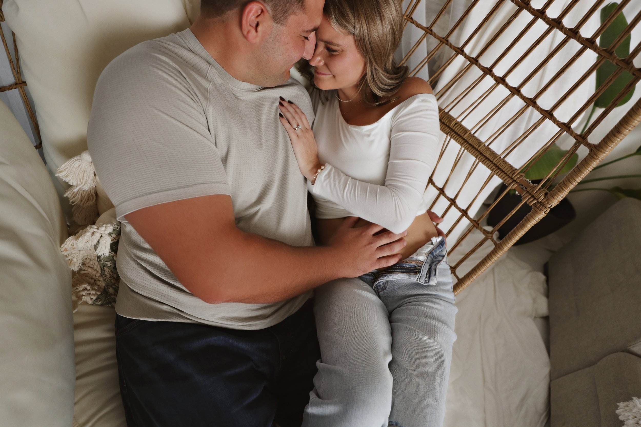 A couple sitting close together on a beige sofa, touching foreheads and smiling, in an intimate, affectionate moment.