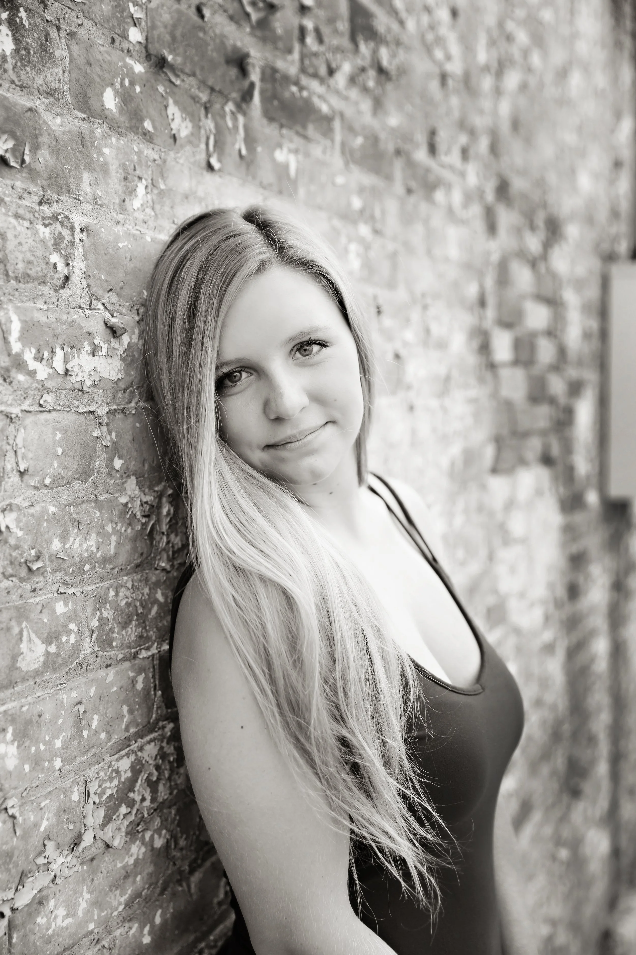 A black and white photo of a young woman with long hair, leaning against a textured brick wall, smiling softly at the camera.