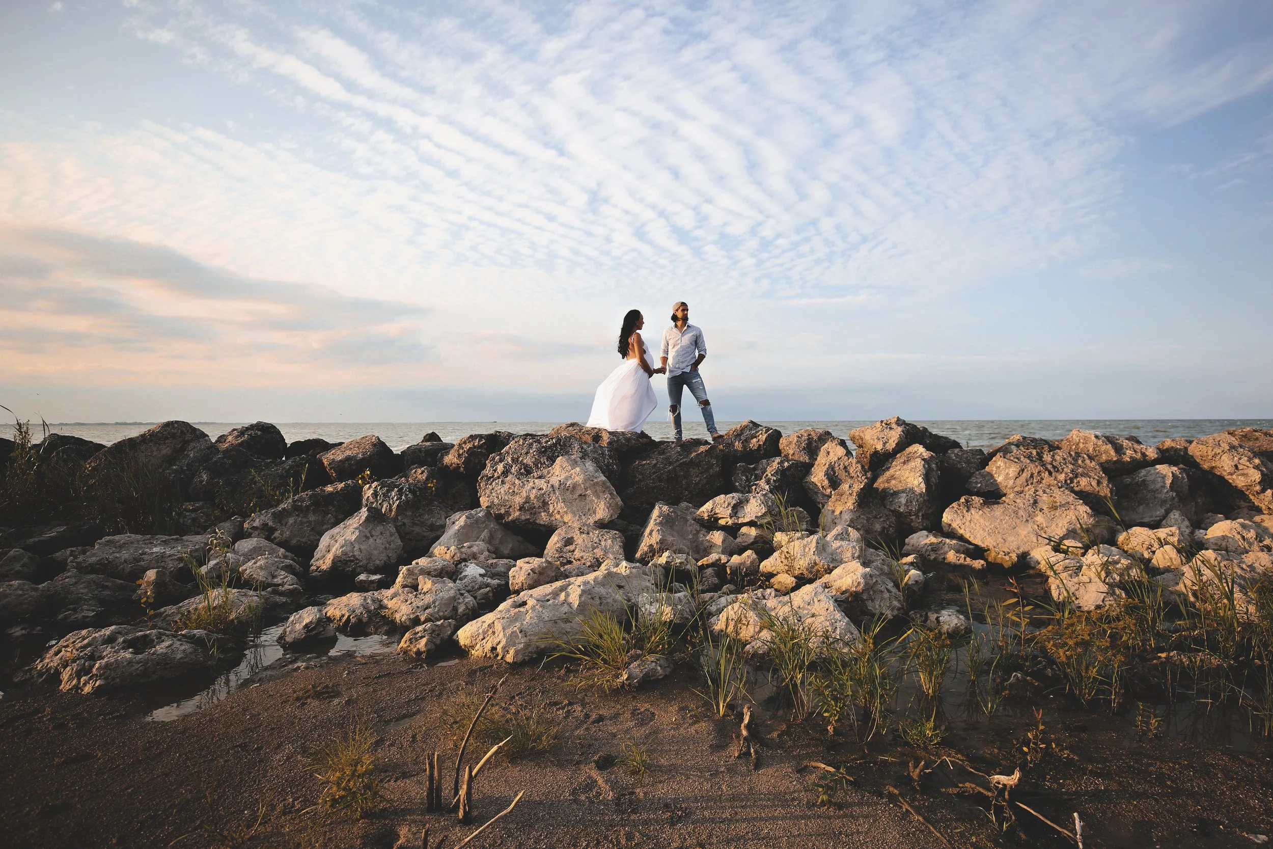 A couple holding hands on rocks at the beach during sunset with a cloudy sky.