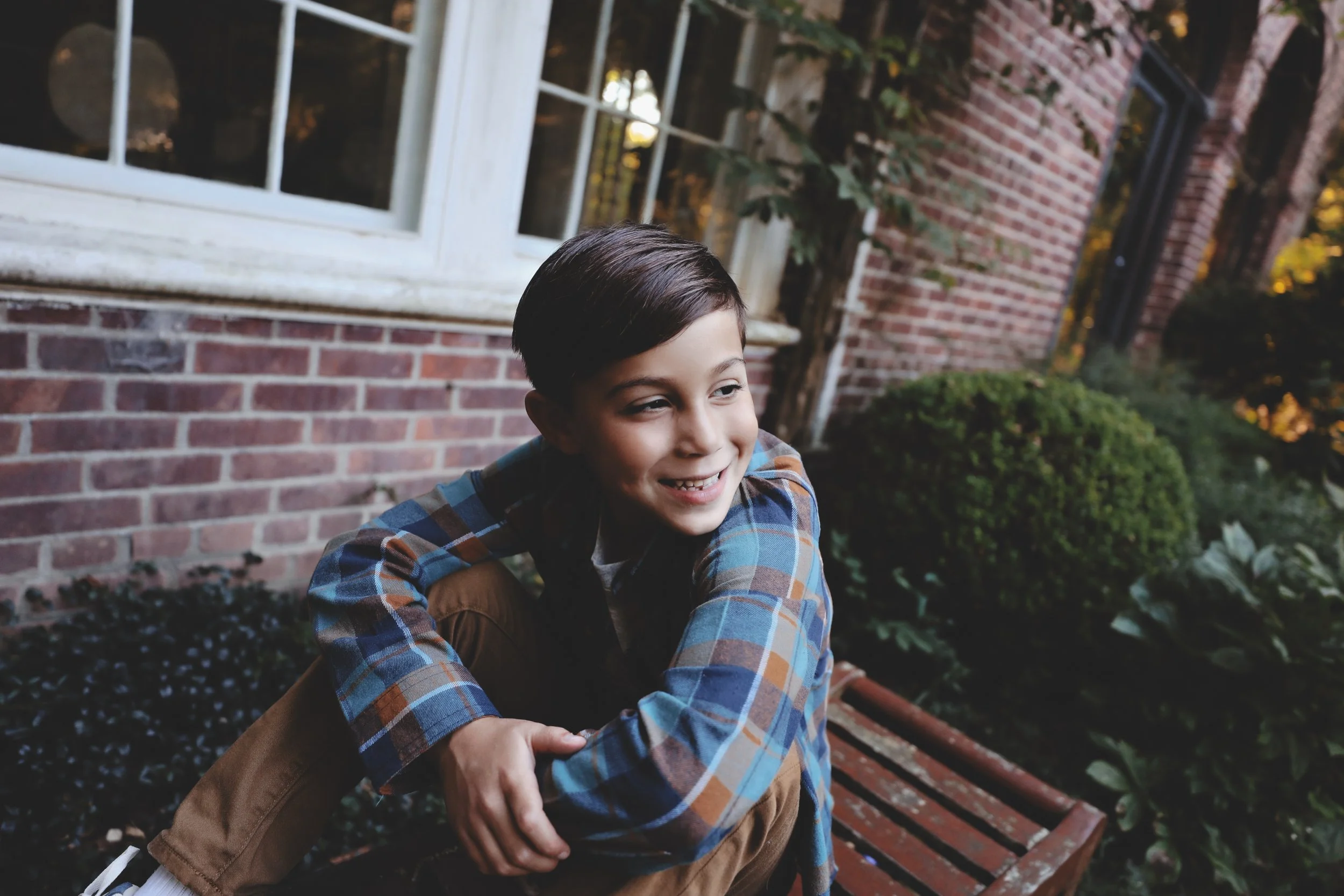 A young boy sitting on a red park bench outside a brick building, smiling and looking to the side.