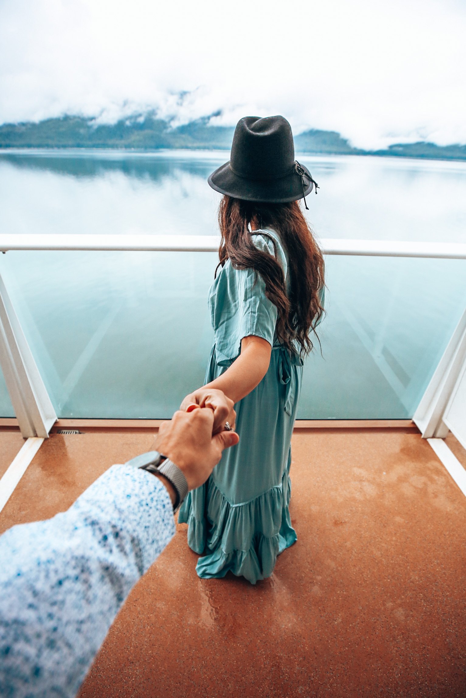 A woman wearing a black hat and turquoise dress holding hands with the photographer while standing on a boat overlooking a calm lake with mountains and mist in the background.