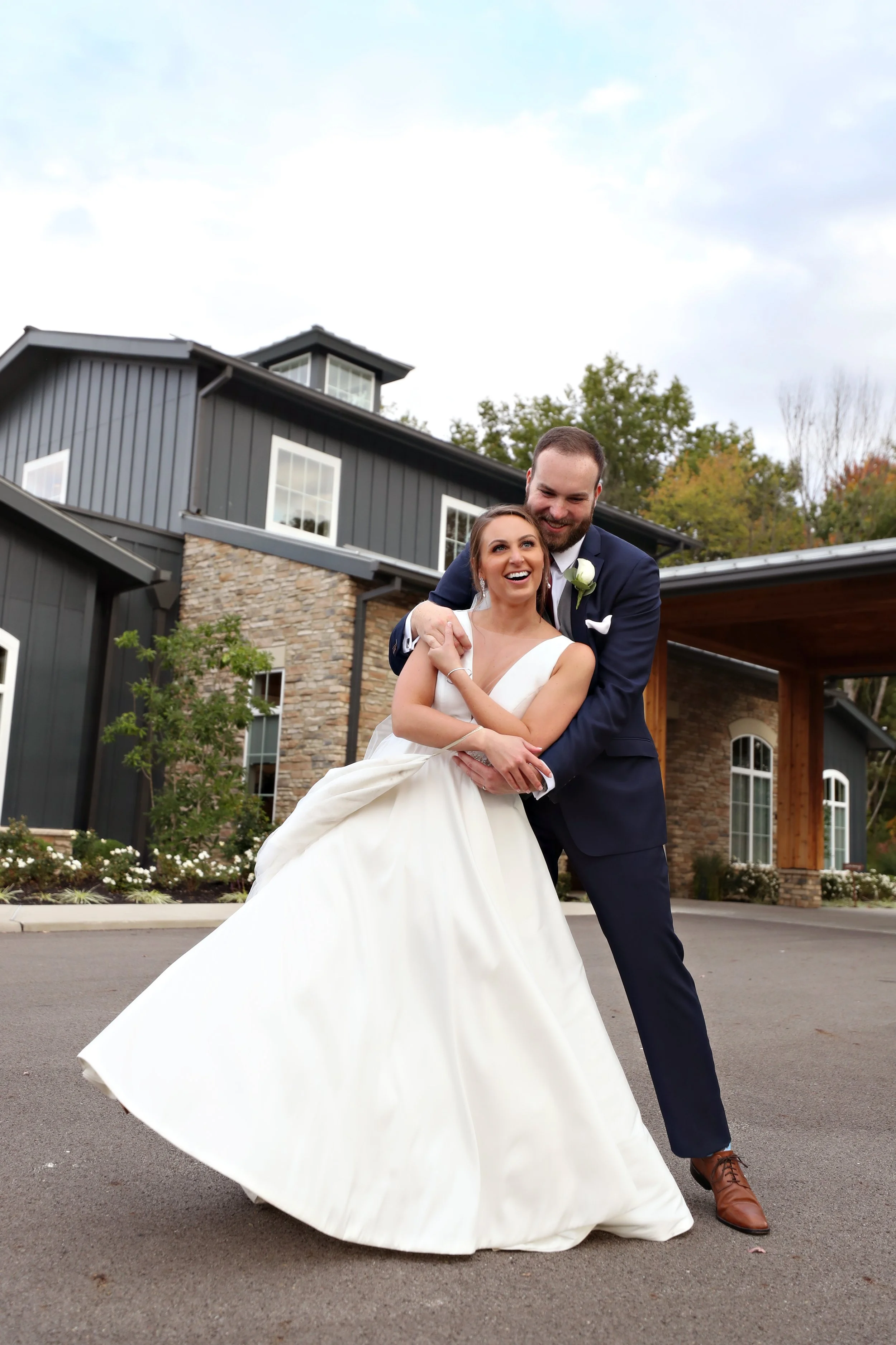 A bride and groom happily dancing outdoors in front of a modern house, with the bride wearing a white wedding dress and the groom in a navy suit.