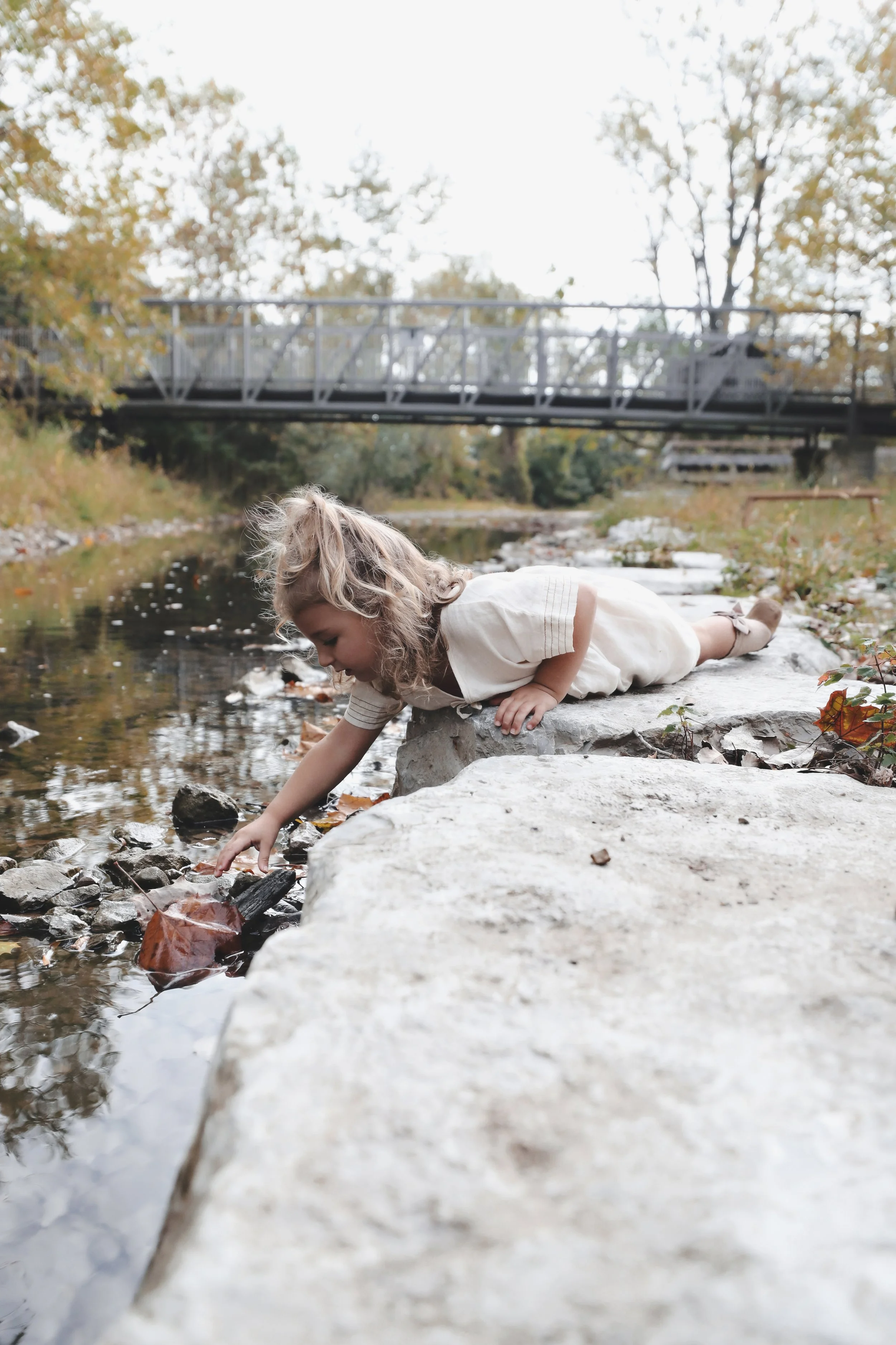 A young girl with curly blonde hair lying on her stomach on a flat rock by a creek, reaching into the water to touch a fallen leaf.