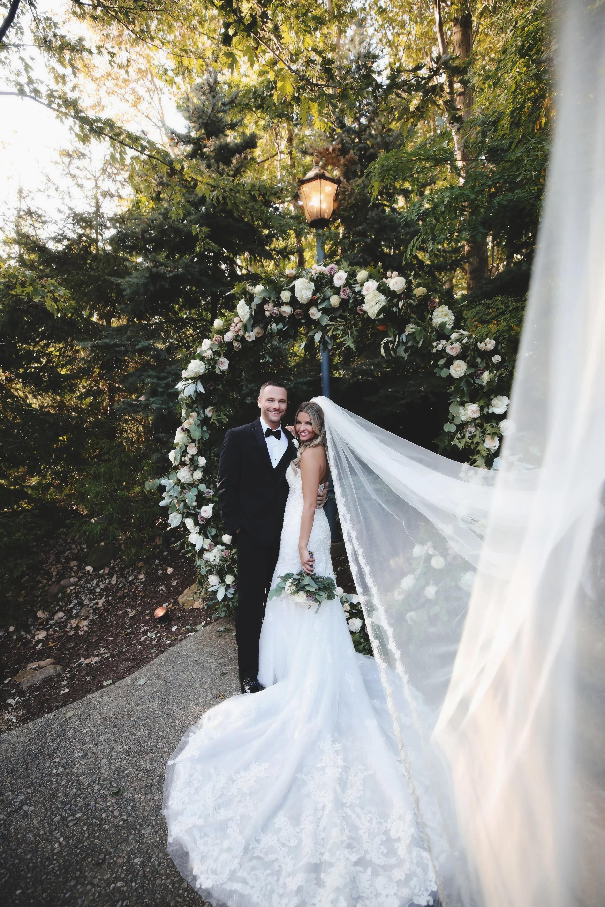 A bride and groom standing together outdoors in front of a floral arch and a street lamp, smiling at the camera on their wedding day. The bride is in a white wedding gown with a long veil, holding a bouquet of flowers. The groom is in a black tuxedo 