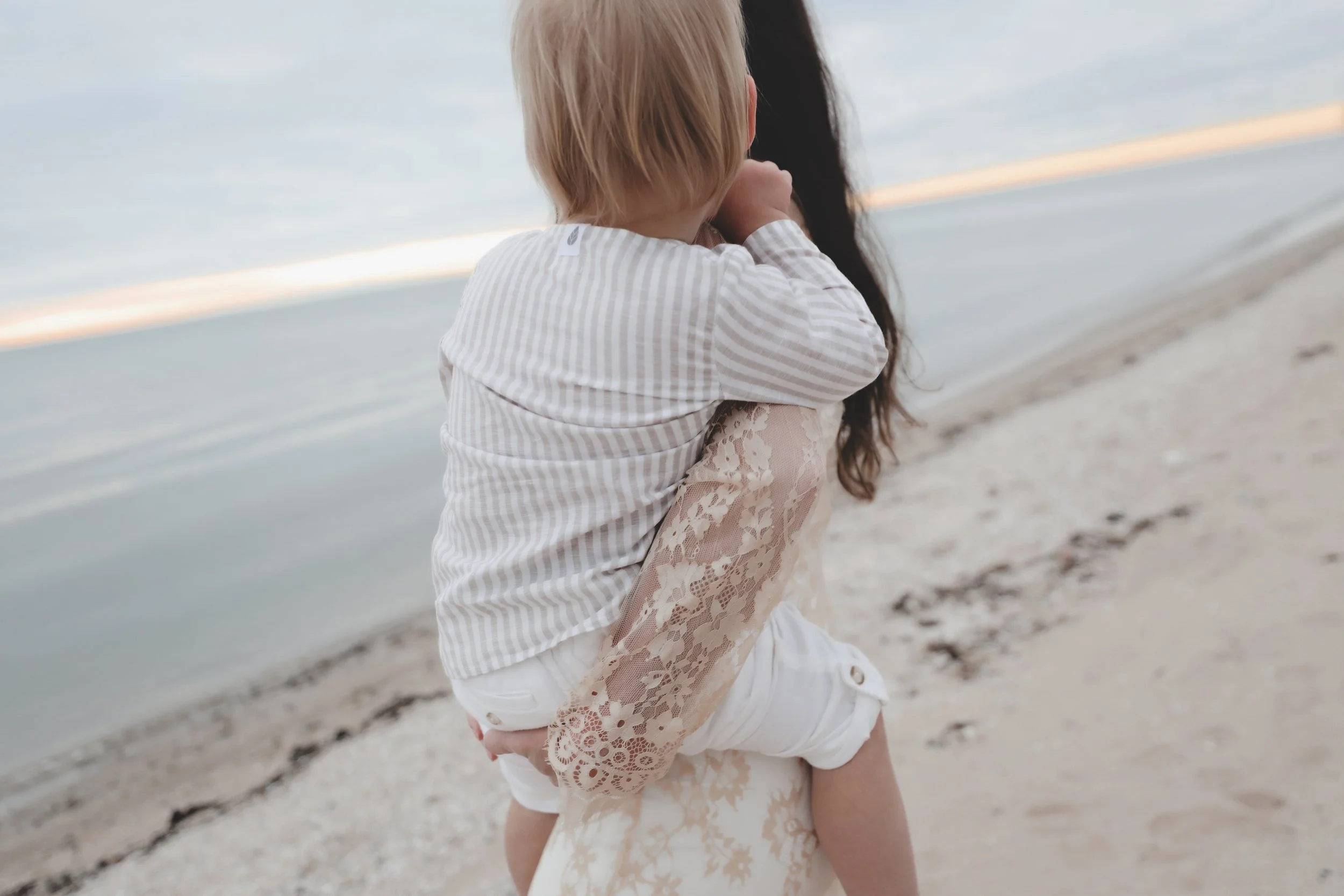 A woman holding a young child on her shoulders at the beach during sunset, with the ocean and cloudy sky in the background.