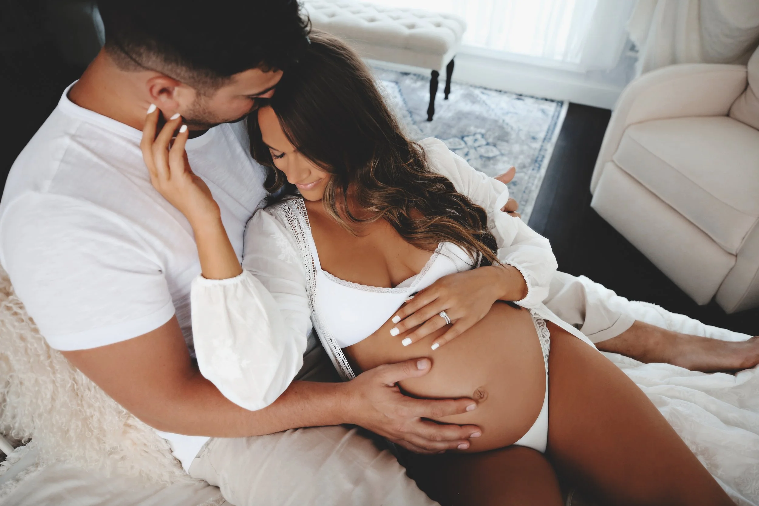 A pregnant woman and her partner in an intimate moment on a bed, with the woman smiling and holding her baby bump, while the partner gently touches her belly.