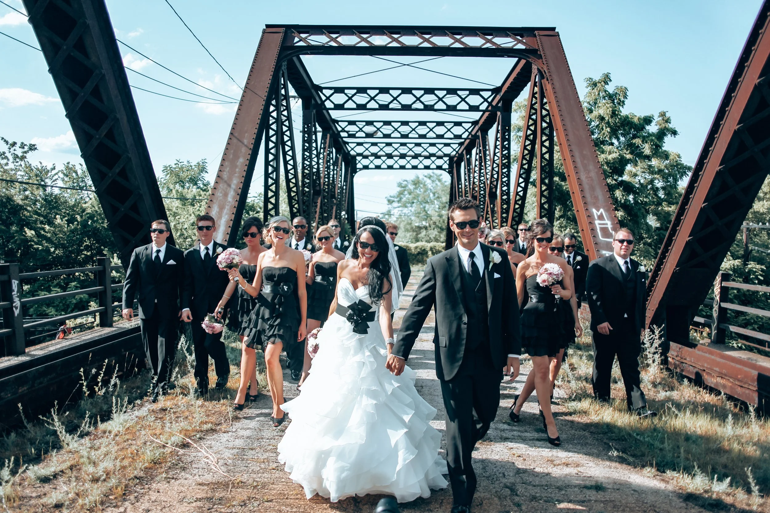 A bride and groom leading a wedding procession of bridesmaids and groomsmen across a rusty iron bridge on a sunny day.