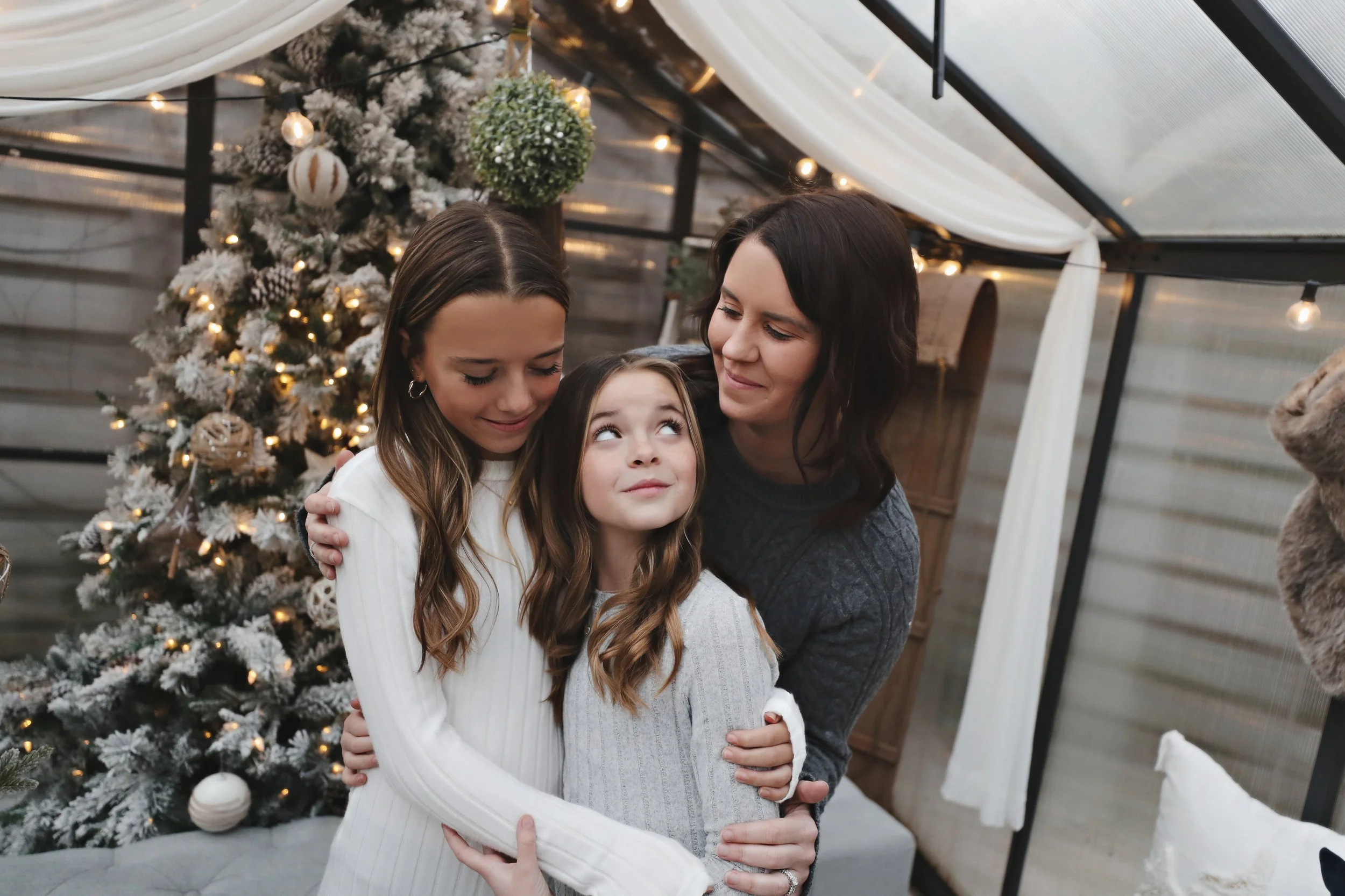 Three people hugging near a decorated Christmas tree, with a cozy indoor setting.