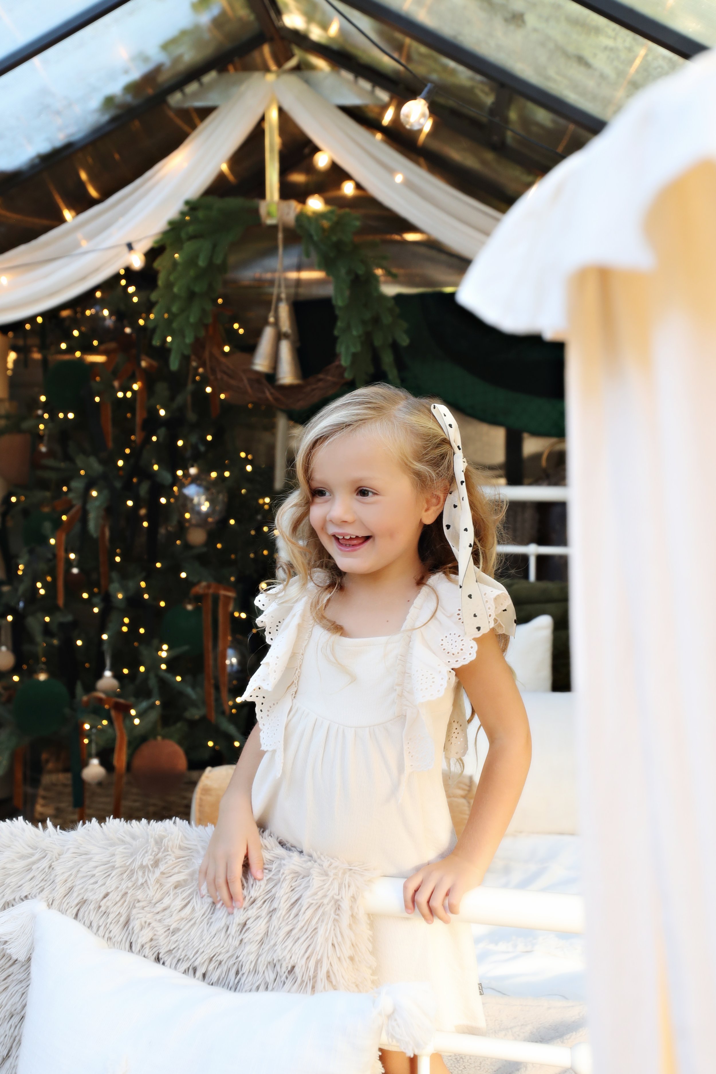 A young girl with blonde curly hair, wearing a white dress with lace sleeves, smiling in a festive decorated space with a Christmas tree in the background.