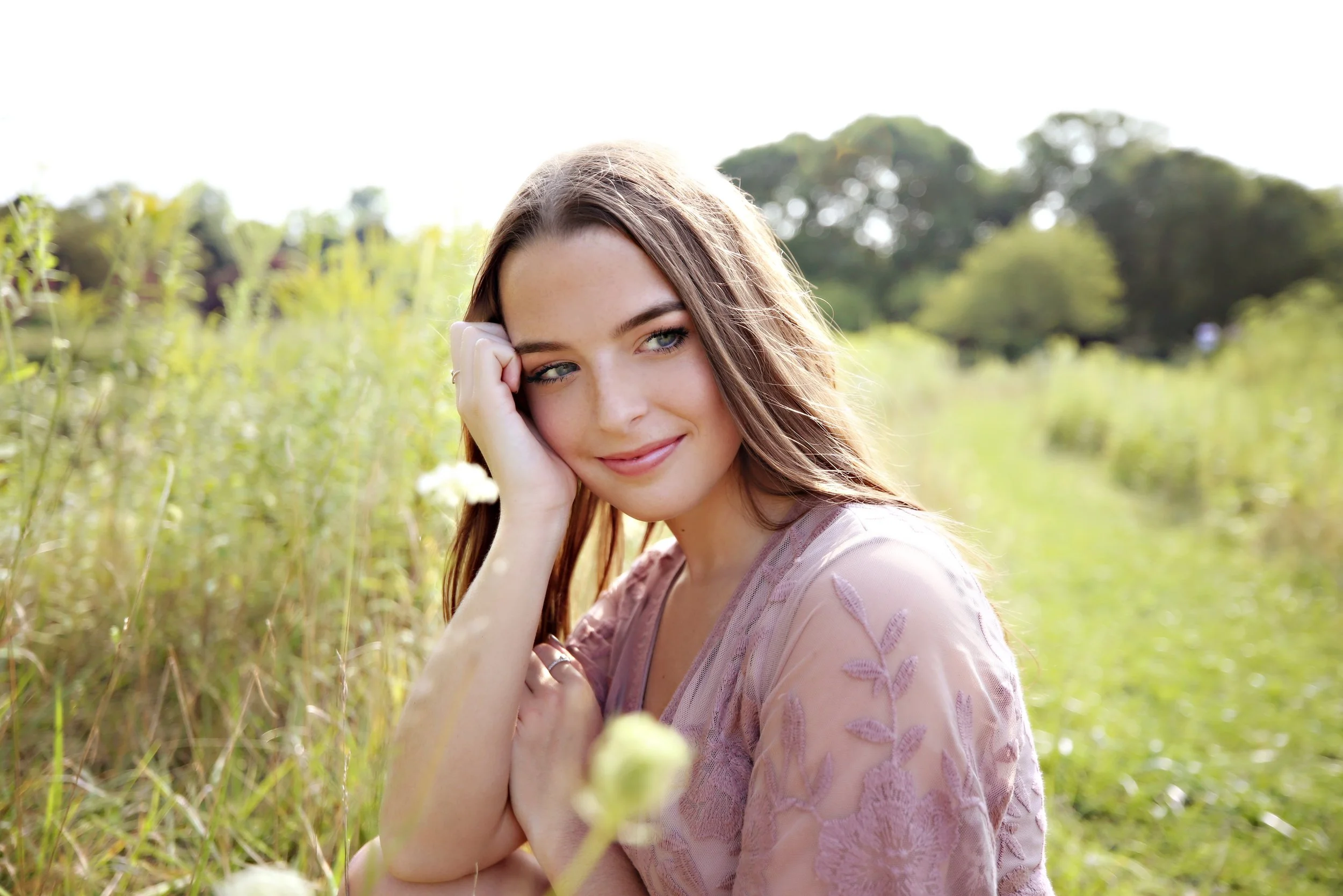 A young woman with long brown hair and fair skin sitting in a field of tall grass with trees in the background, smiling softly at the camera.