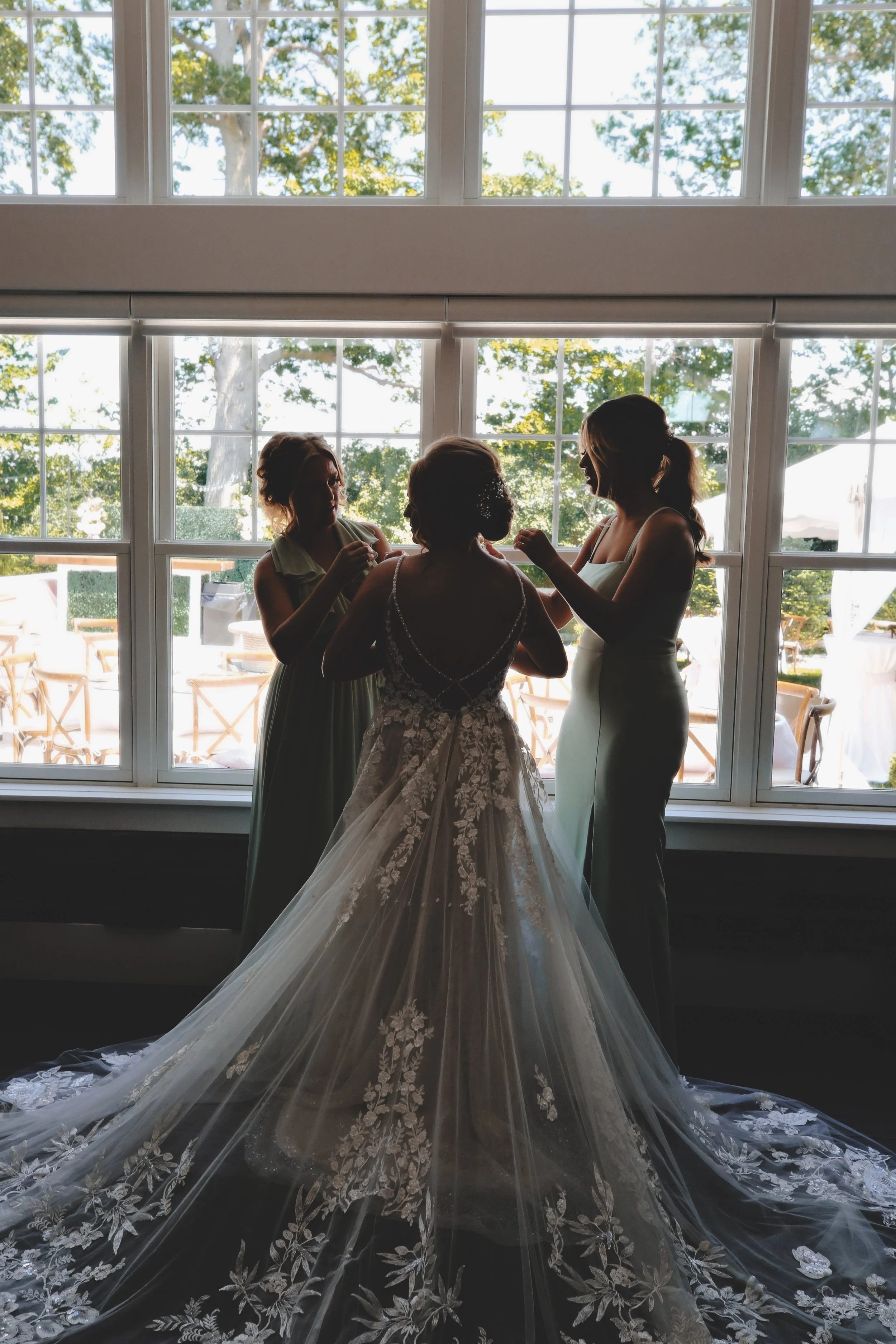 Silhouette of a bride in a wedding gown and two bridesmaids adjusting her dress by a large window, outdoor scene with trees and chairs visible outside.