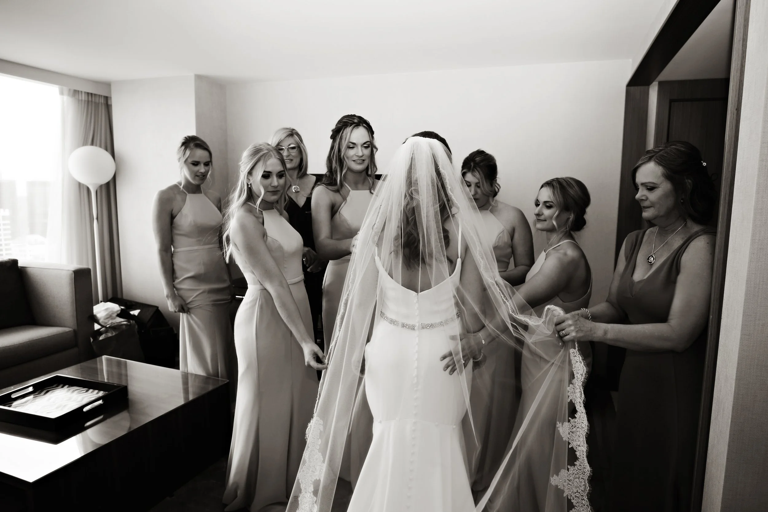 A bride in a wedding dress adjusting her veil with her bridesmaids and women helping her.