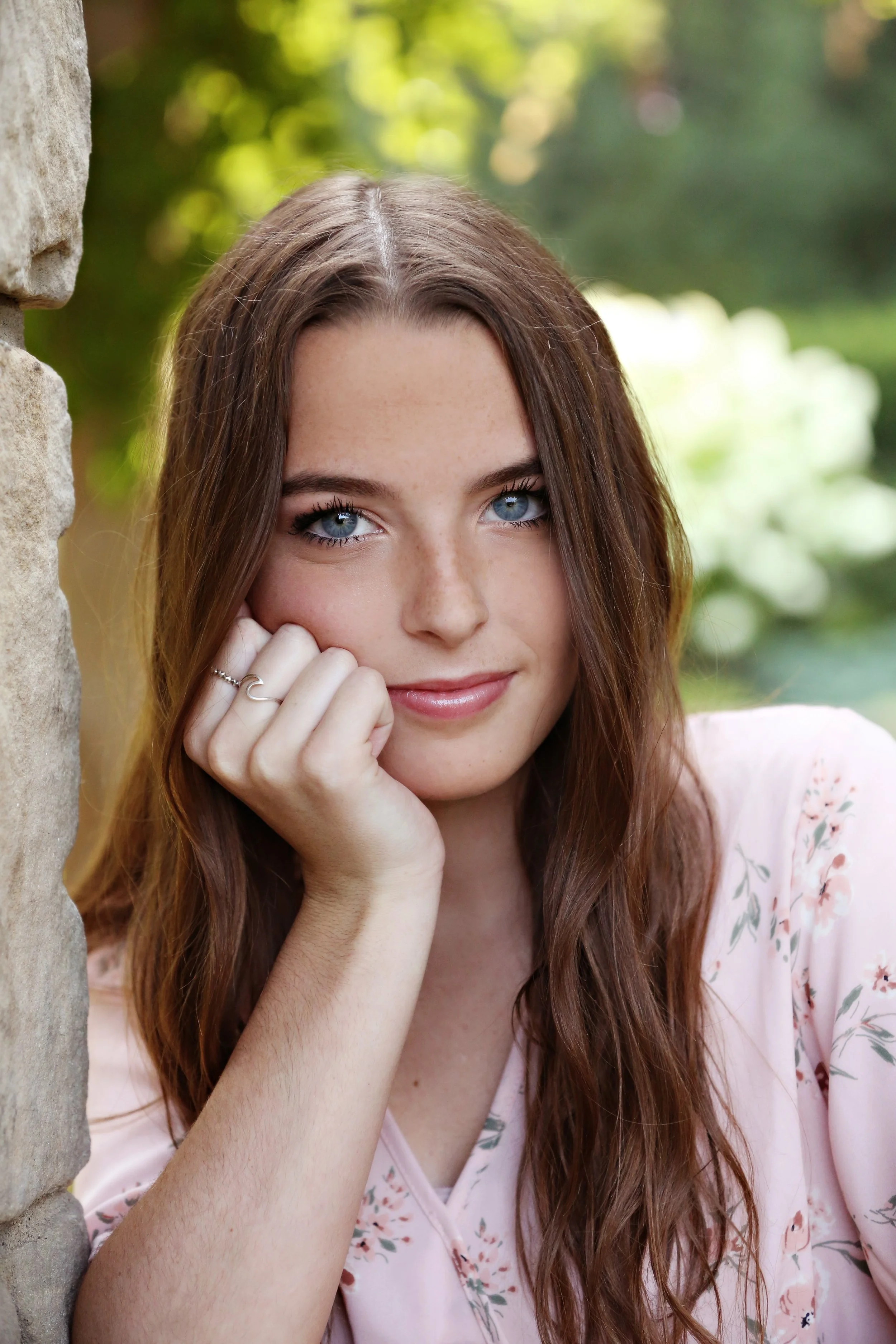 Close-up of a young woman with long brown hair and blue eyes, resting her face on her hand next to a stone wall