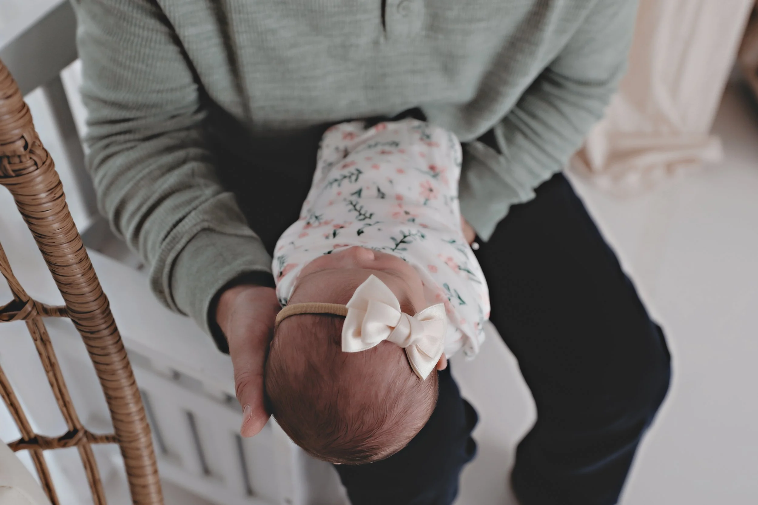 Person holding a newborn baby dressed in floral clothing with a cream bow headband, sitting on a white crib.