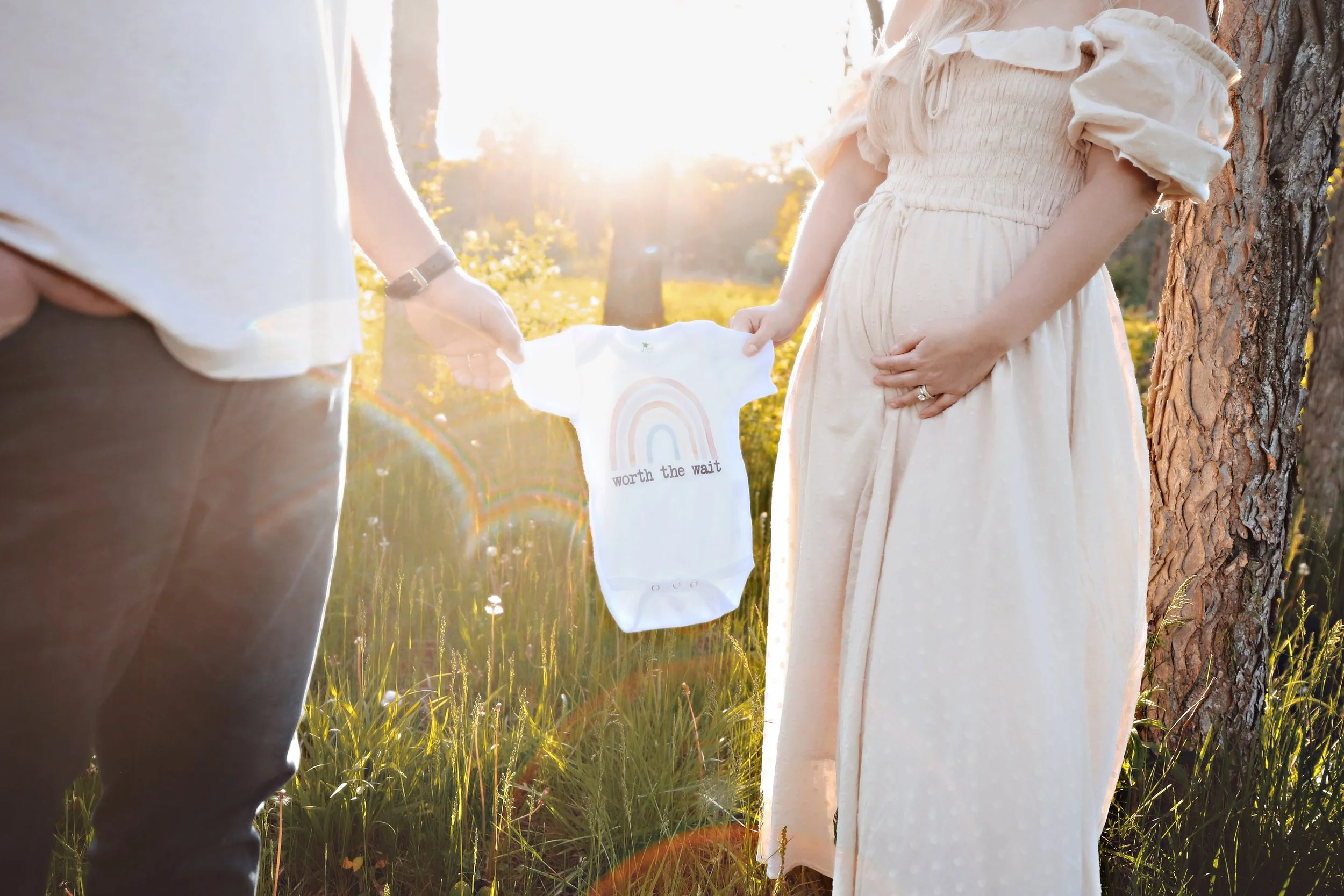 A pregnant woman and a man holding an ultrasound baby onesie outdoors during sunset, with grass and trees in the background.