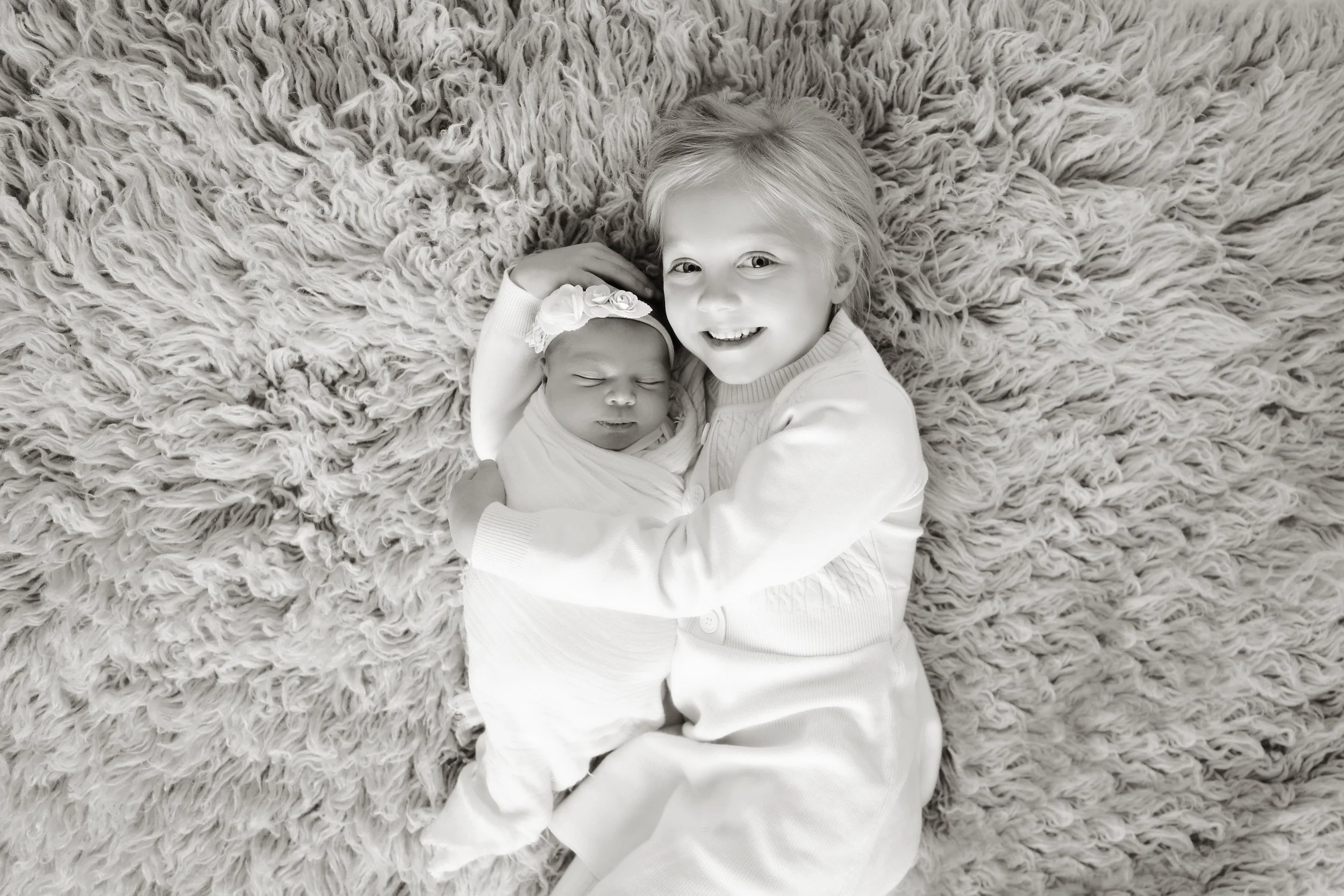 A young girl and a newborn baby girl lying on a fluffy rug. The older girl is smiling at the camera and gently holding the baby, who is sleeping with a headband and a floral decoration.