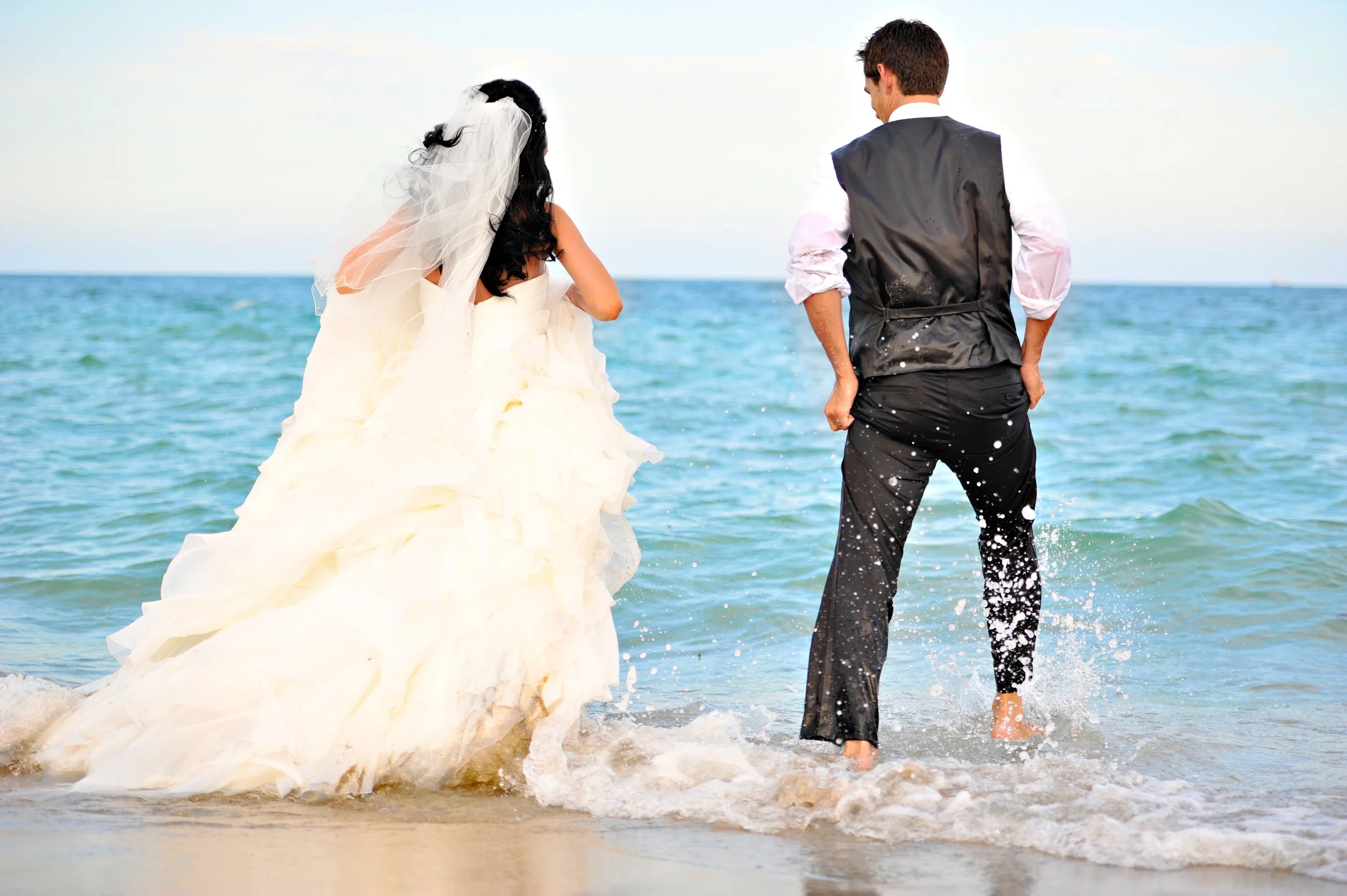 Bride and groom standing in the water at the beach, facing the ocean, with waves around their feet during their wedding.