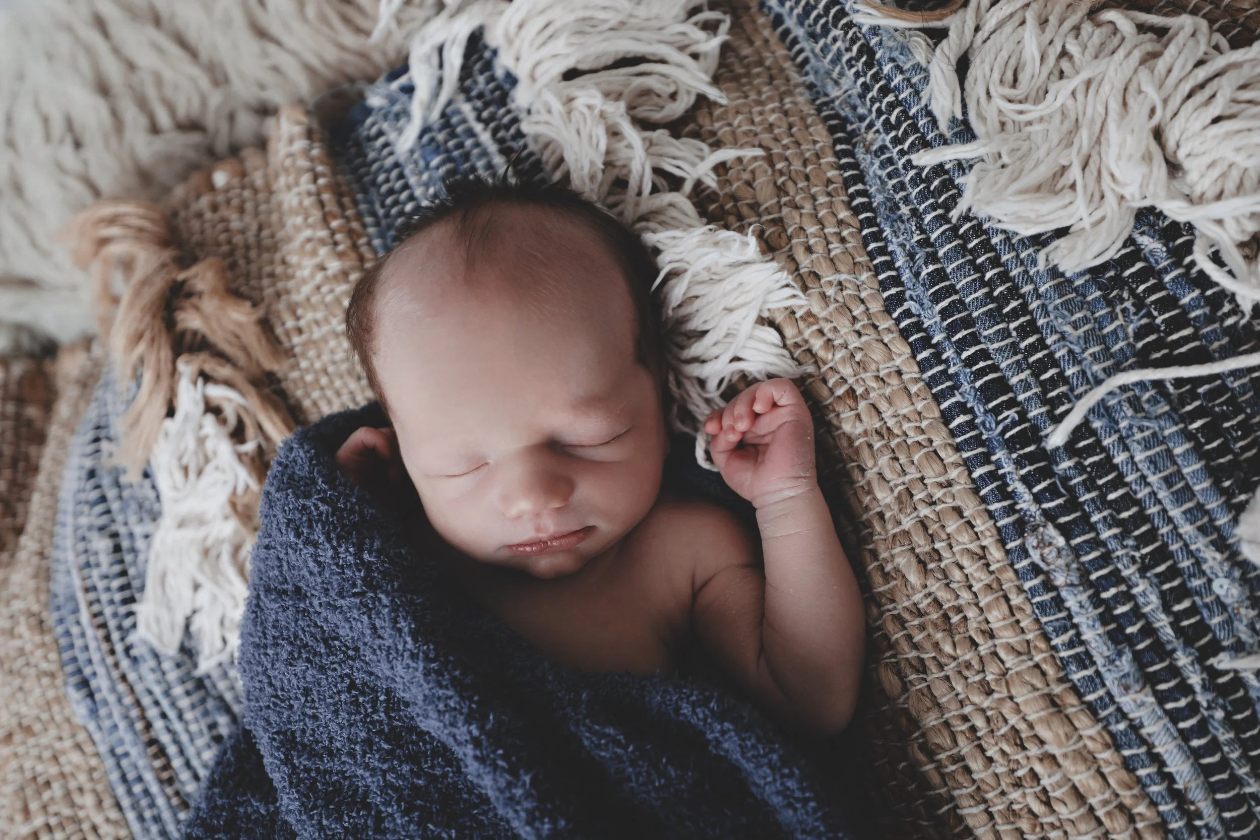 A sleeping newborn baby wrapped in a dark blue towel, lying on a woven blanket with white fringes.
