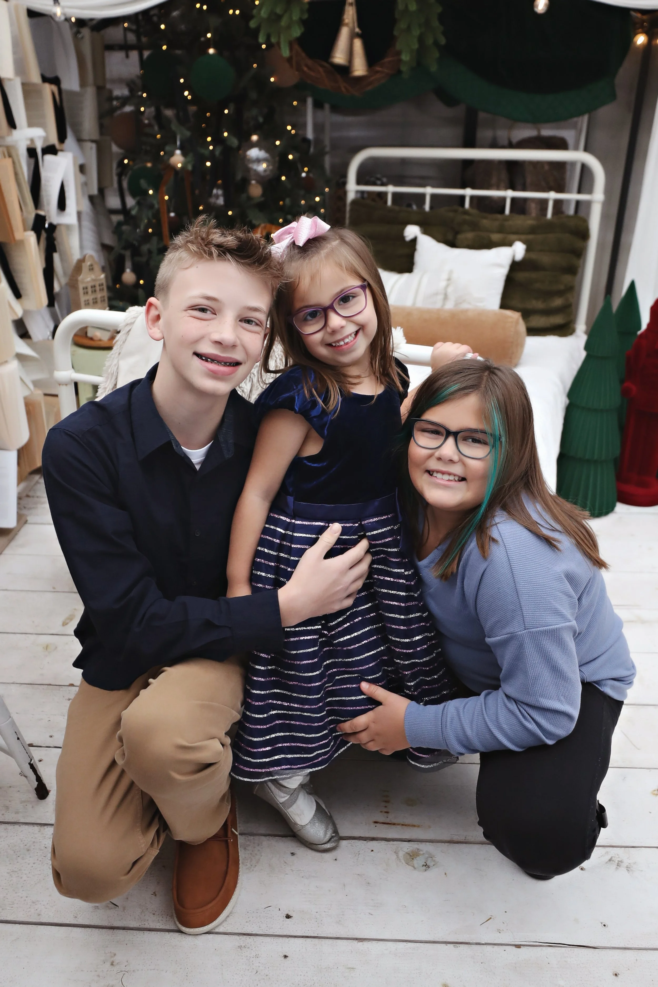 Three children smiling and posing together in front of a Christmas decorated background, including a Christmas tree with lights and ornaments, and holiday-themed decorations.