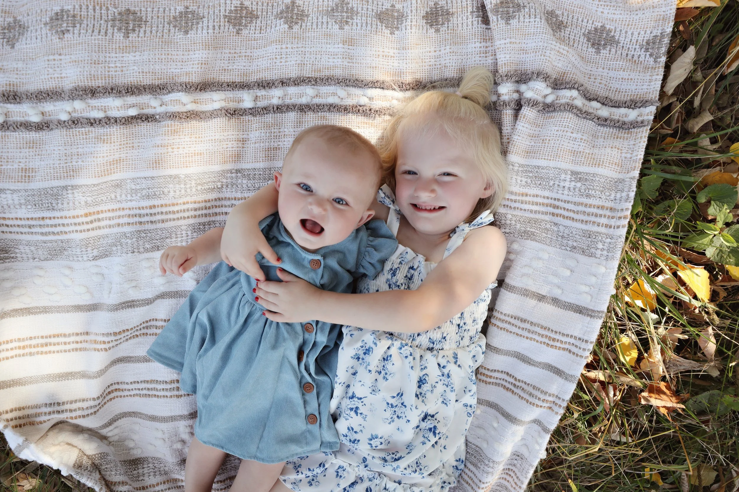 A young girl and a baby lying on a striped blanket outdoors, smiling and hugging each other.