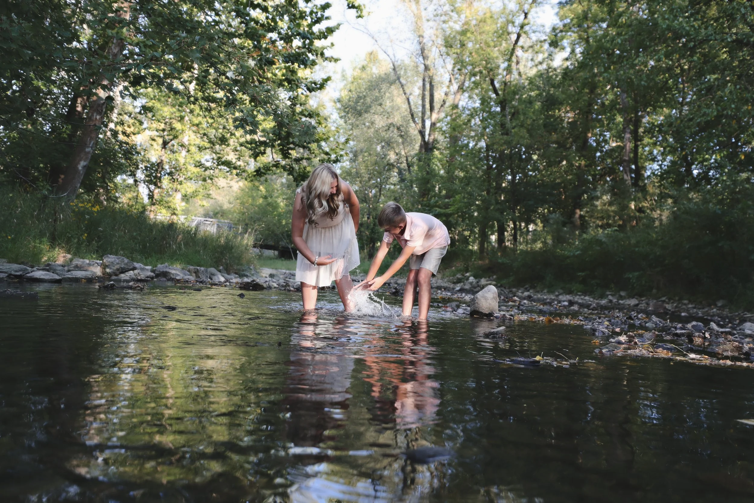 A woman and a boy playing and splashing water in a creek in a forested area during daytime.