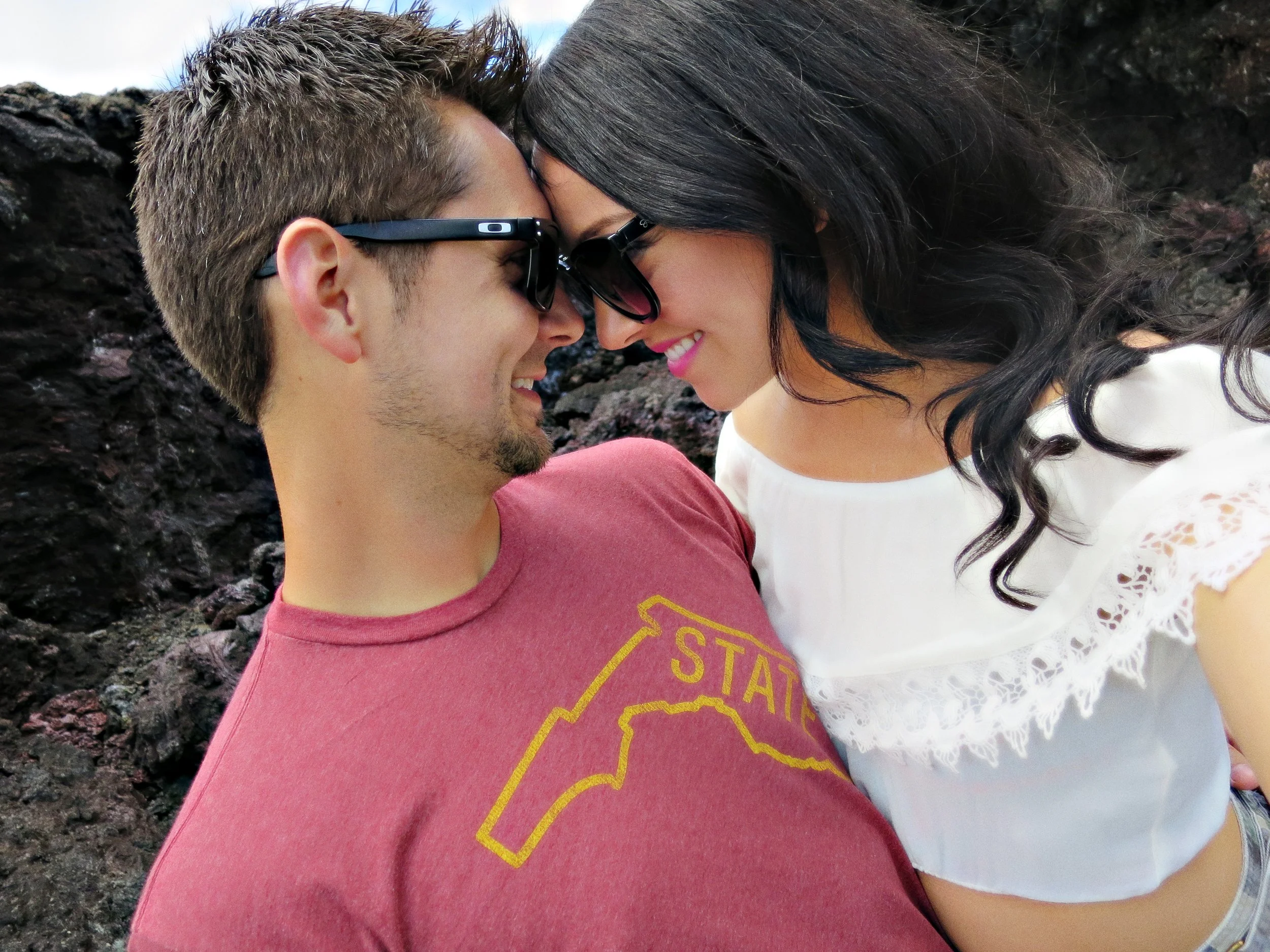 A smiling young couple is close together, touching foreheads, both wearing sunglasses, outdoors near rocky terrain.