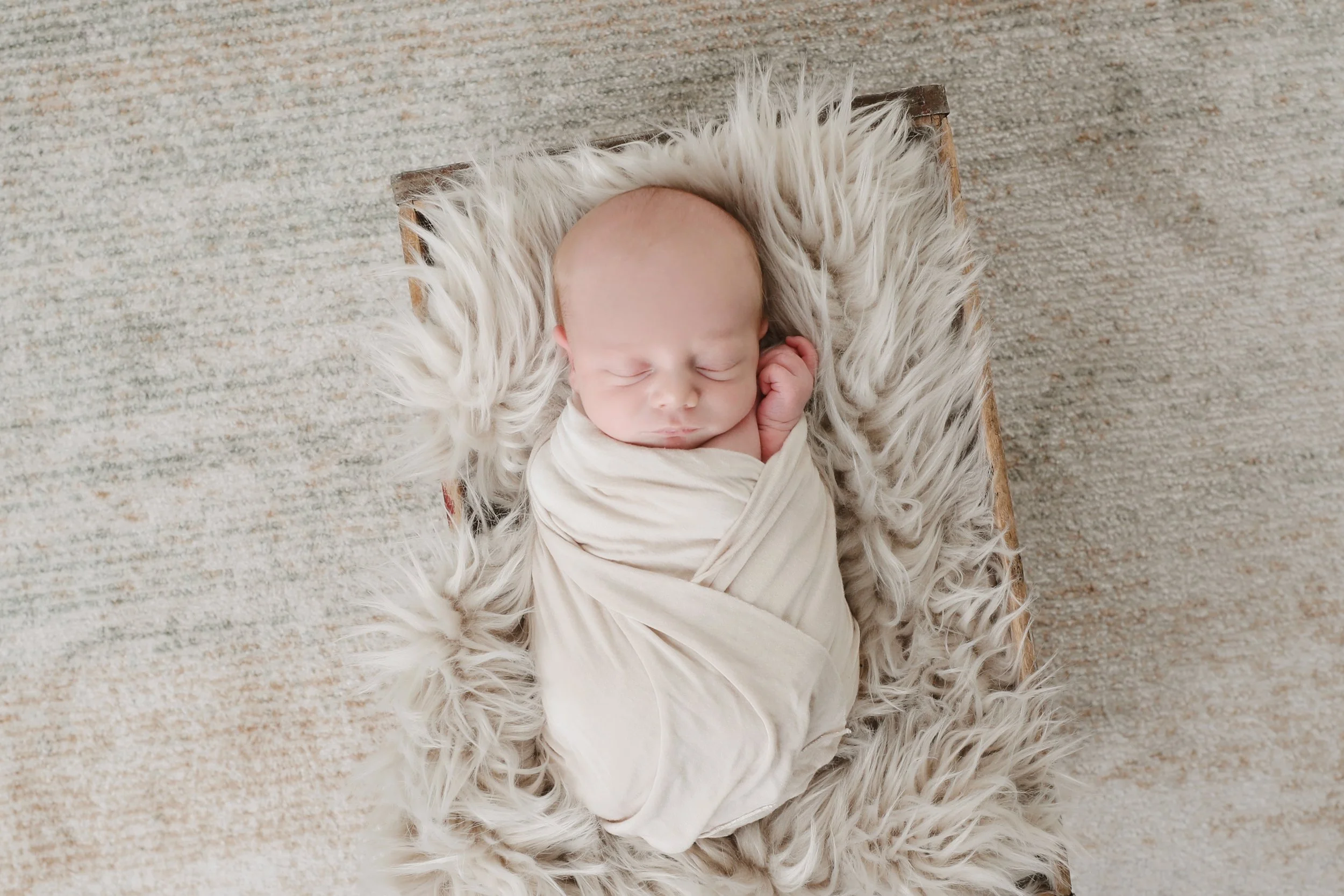 A sleeping baby wrapped in a cream-colored blanket, lying on a soft, fluffy white rug on a wooden bed.
