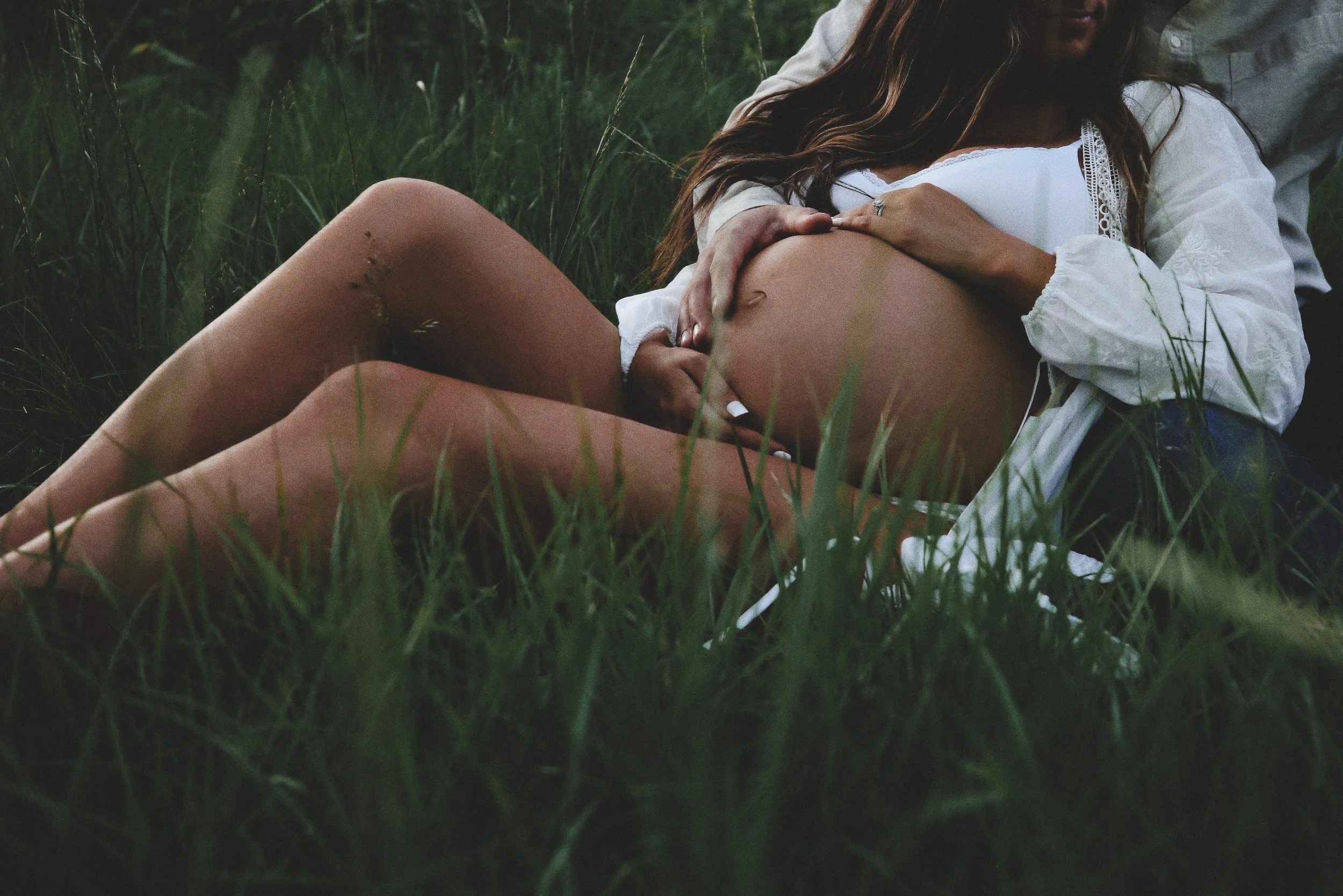 A pregnant woman lying on her back in tall grass with a man behind her, both partially visible, during twilight or dusk.