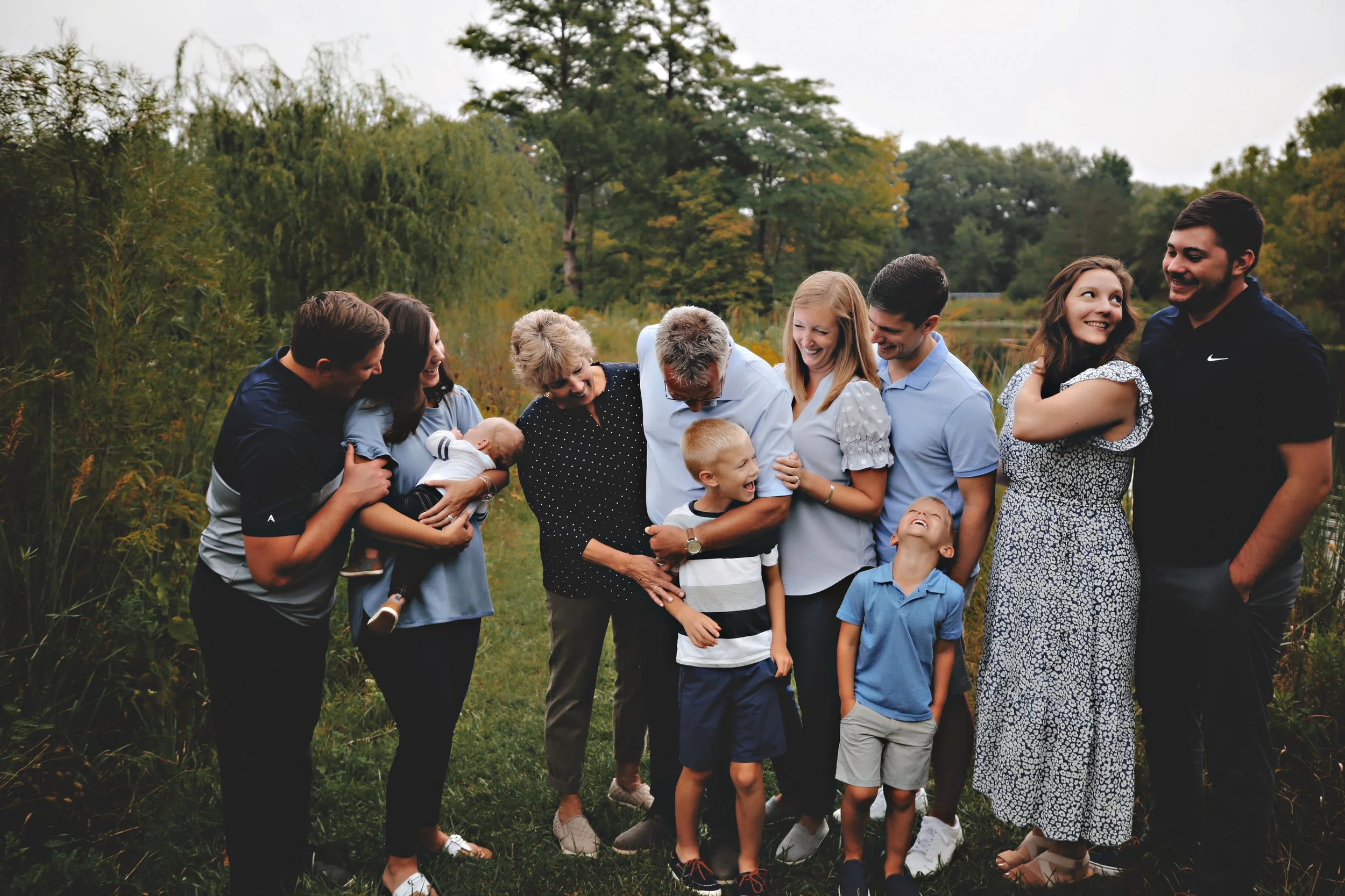 A multi-generational family standing outdoors on a grassy area by a lake, laughing and embracing each other.