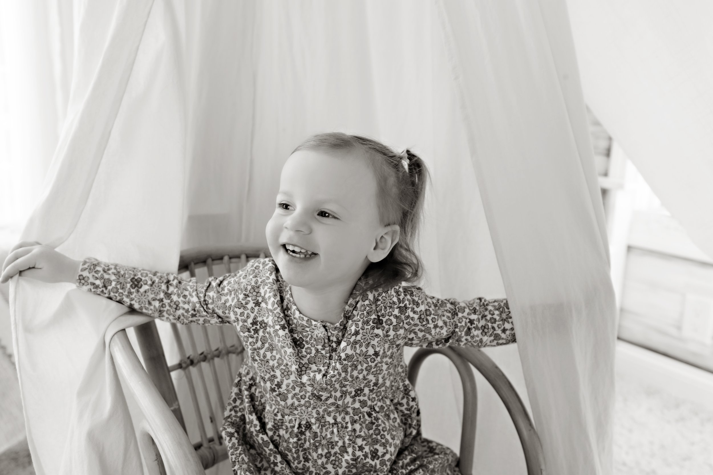 A young girl with a ponytail and a floral dress sitting in a wicker chair, smiling and holding onto a fabric canopy around her. The photo is in black and white.