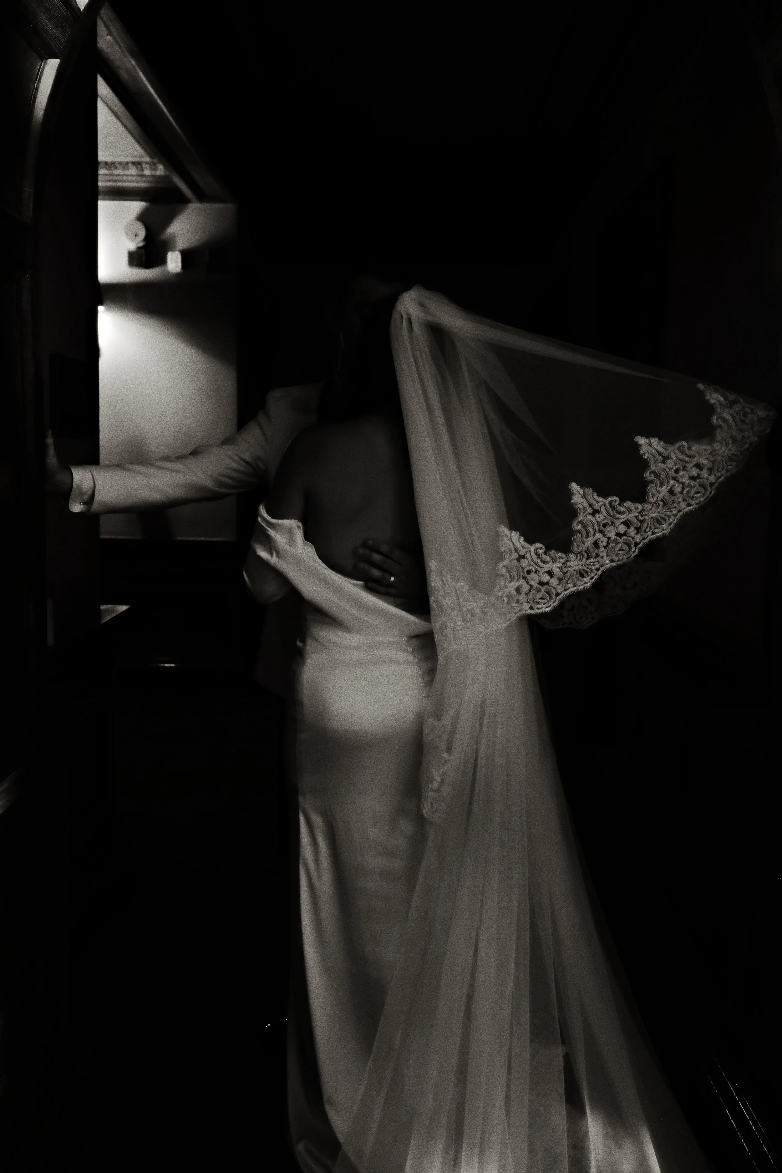 A bride in a wedding dress with an off-shoulder design and a long veil, standing close to a person in a suit who is helping her with her dress or preparing her for her wedding.