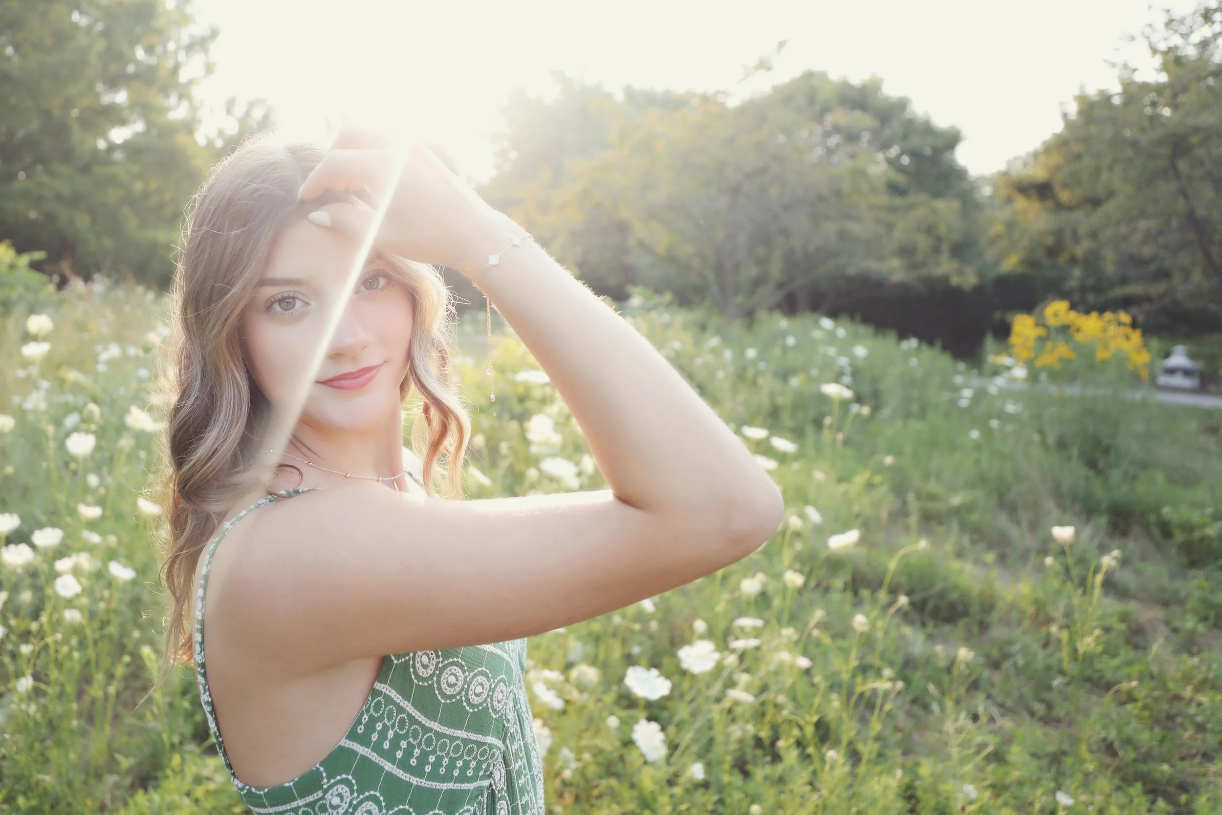 Young woman with wavy hair in a green patterned dress standing in a field of white flowers with trees in the background, sunlight shining over her head.