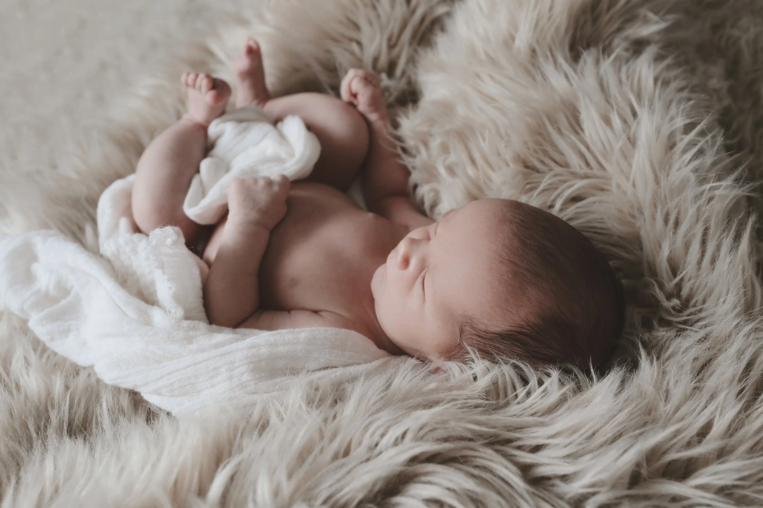 A newborn baby lying on a soft, fluffy surface, wrapped in a white cloth and holding a small cloth, resting peacefully with eyes closed.