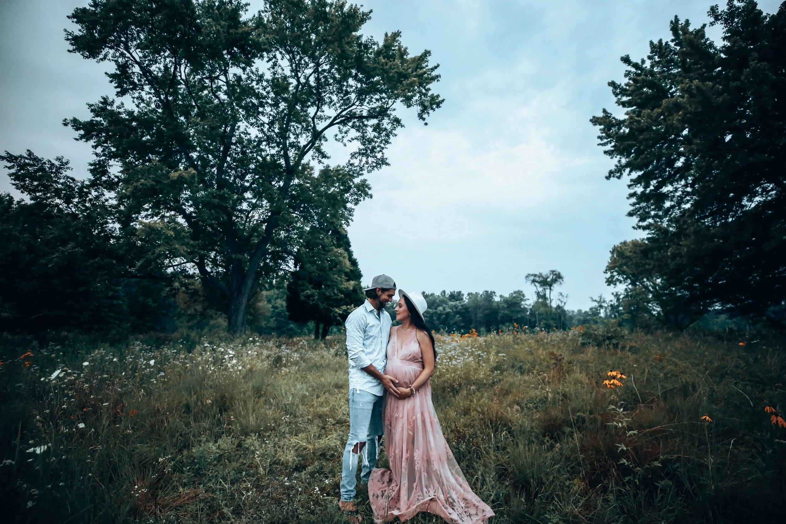 A couple standing close together in a grassy field with flowers, surrounded by trees and a cloudy sky.