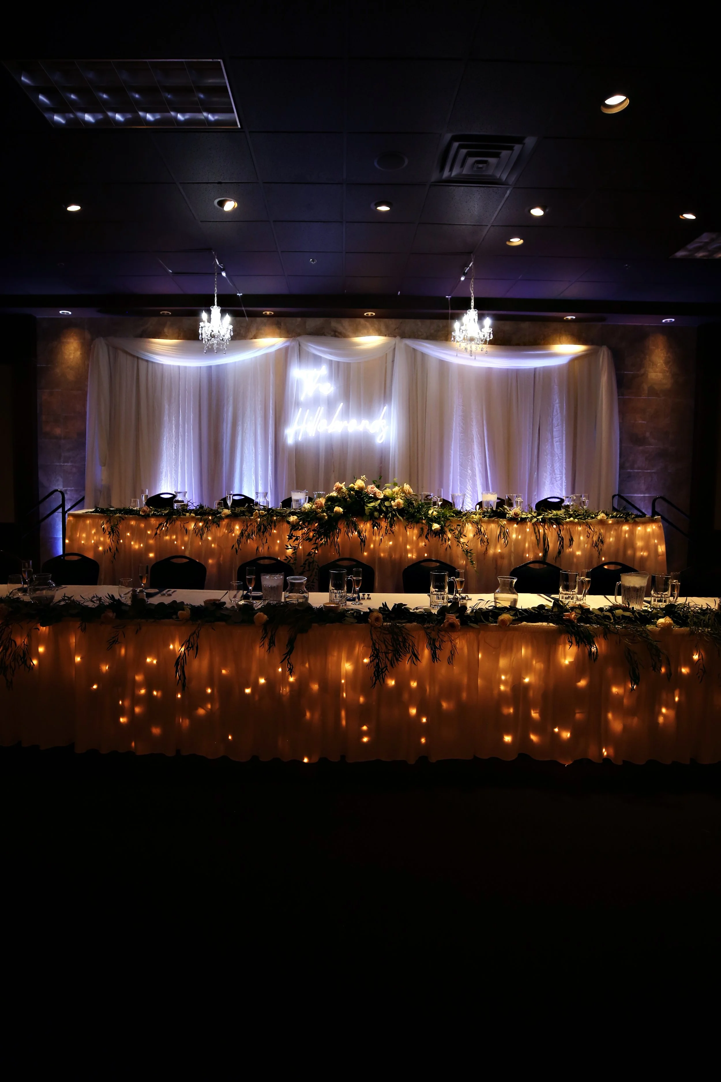 Decorated event reception table with flowers, candles, and string lights, illuminated by chandeliers, with a white draped backdrop featuring neon sign reading 'Love & Happiness'