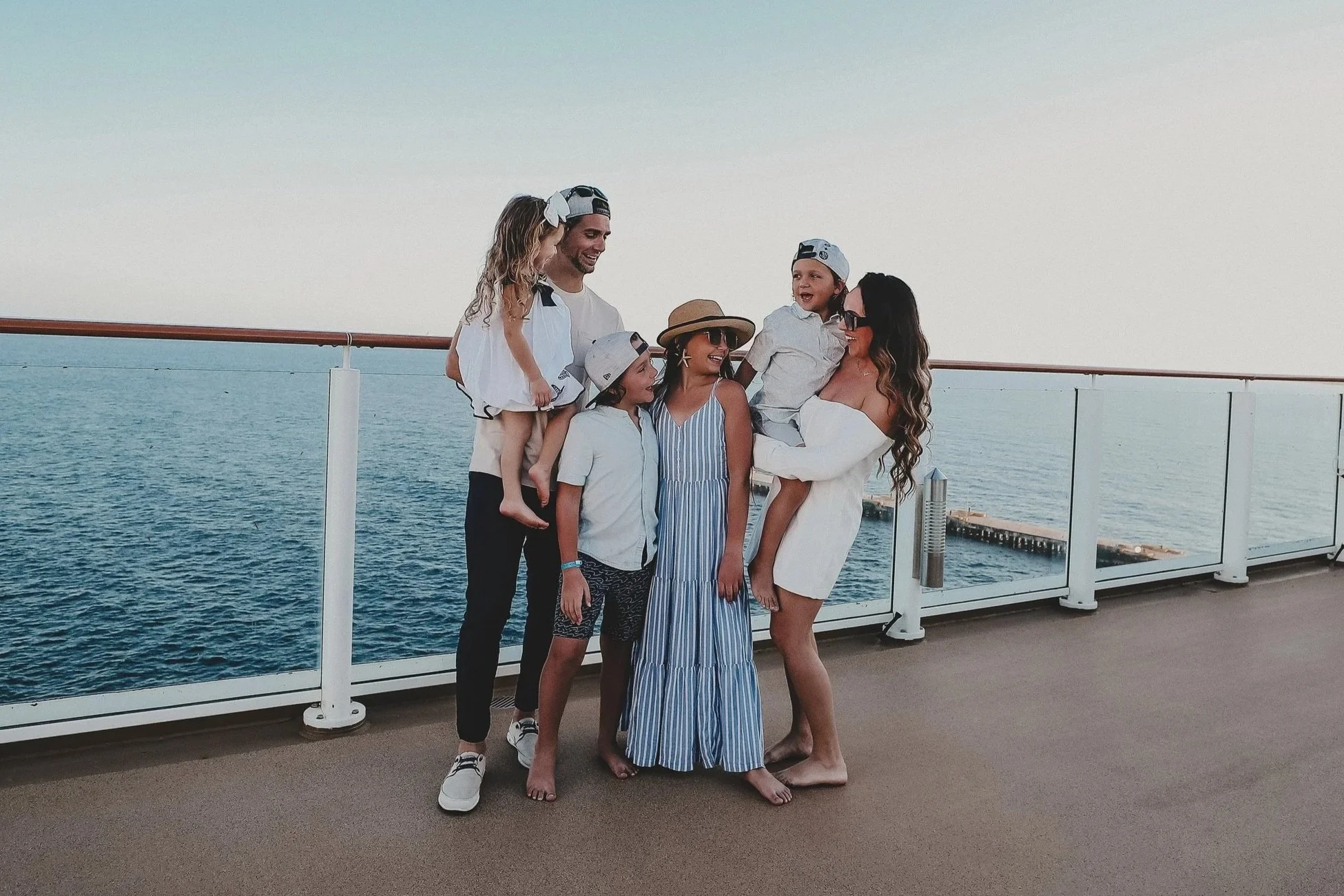 A family of six enjoying a day at sea on a boat deck, with ocean and sky in the background. They are smiling and interacting with each other, dressed in casual, summer clothing.