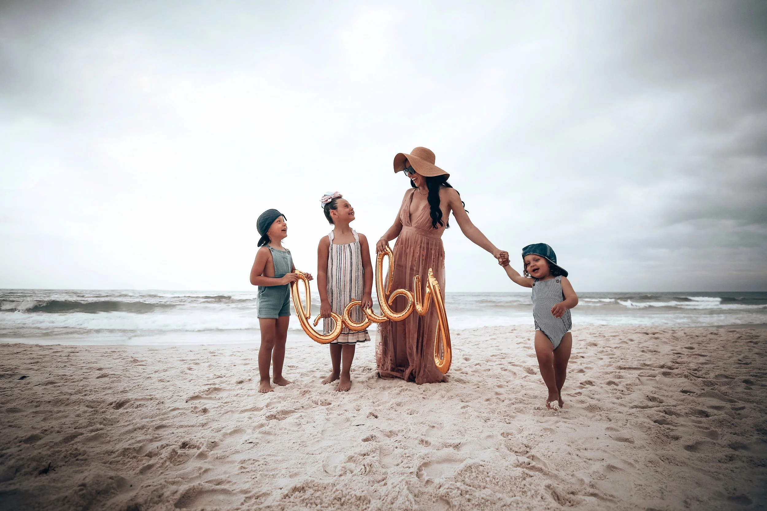 A woman and three young girls on a sandy beach holding a balloon spelling 'baby' with ocean waves in the background.