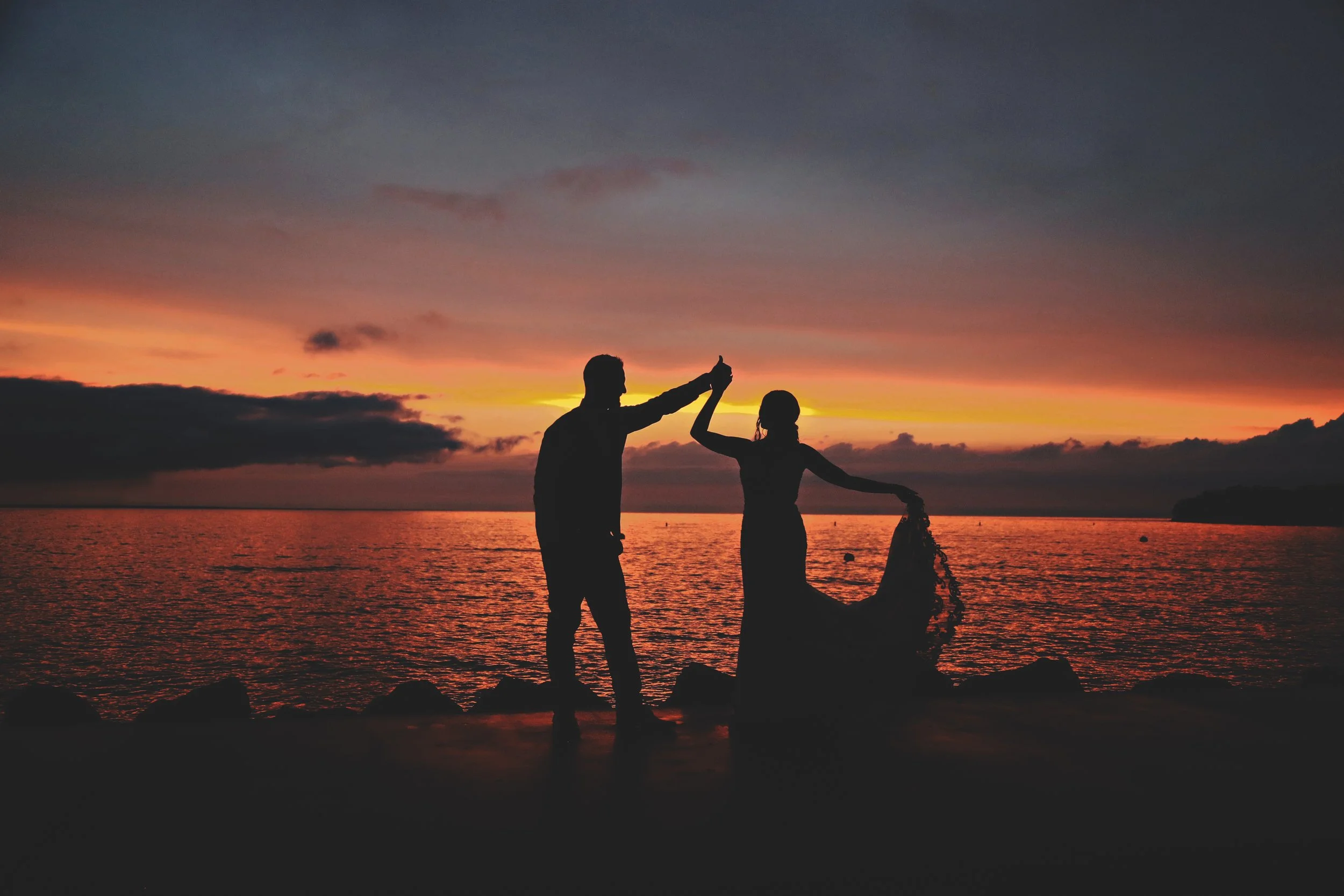 Silhouettes of a man and woman dancing on a rocky shore during sunset over the ocean.