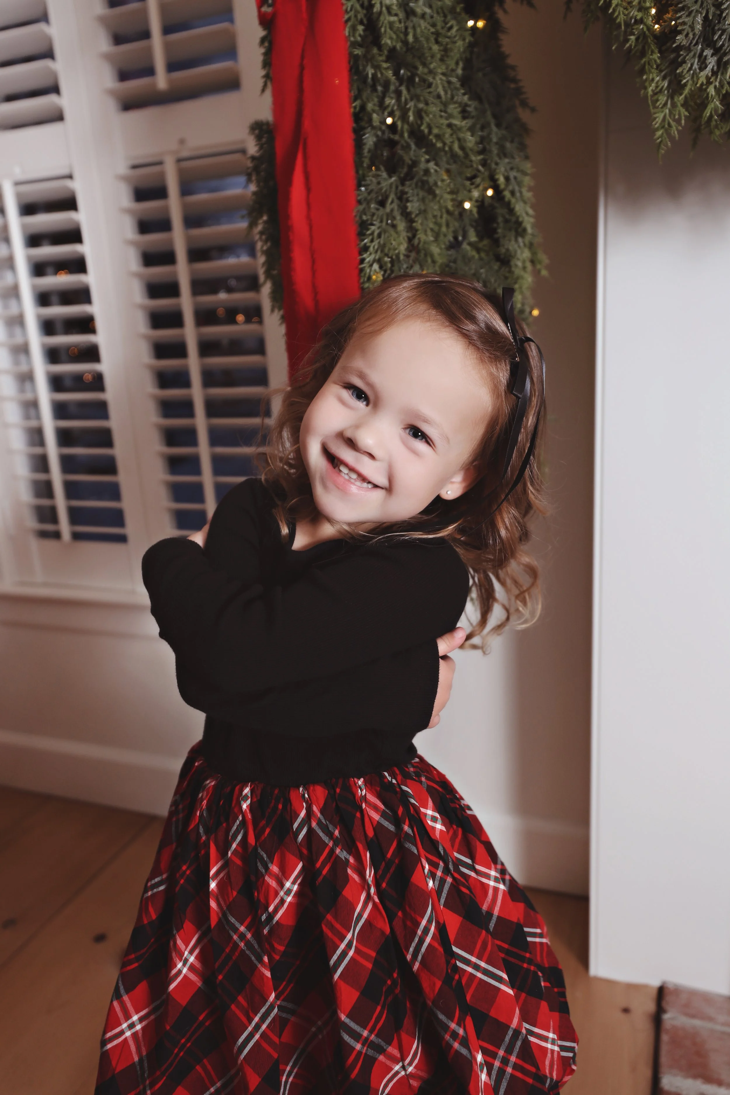 A smiling young girl with curly hair, wearing a black top and a red plaid skirt, standing indoors beside a Christmas tree with green branches and red ribbon decorations.