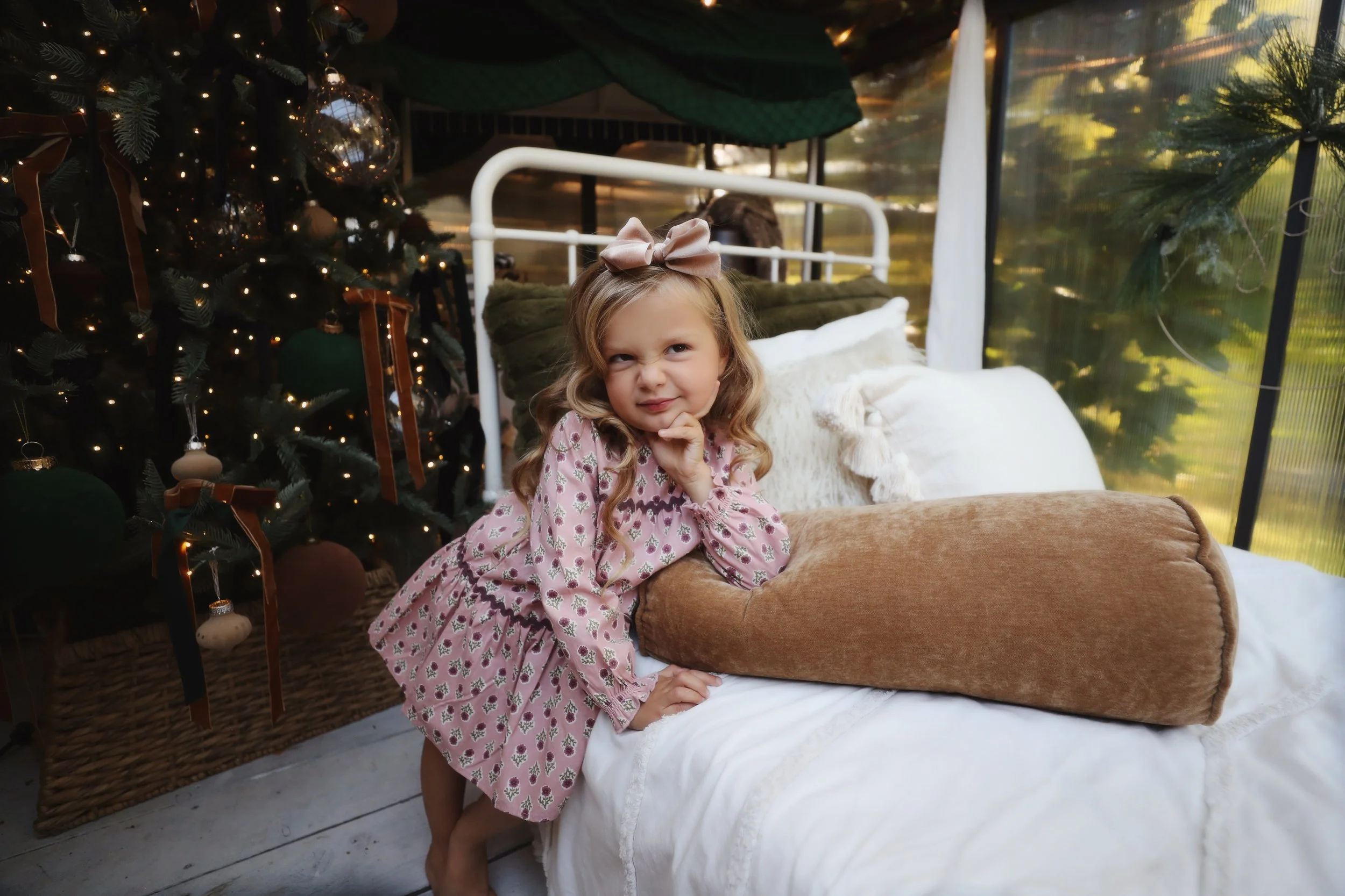 A young girl with long wavy hair, wearing a pink dress with a floral pattern and a pink bow headband, standing on a sofa with decorative pillows, beside a decorated Christmas tree with ornaments and lights, in a sunlit room.