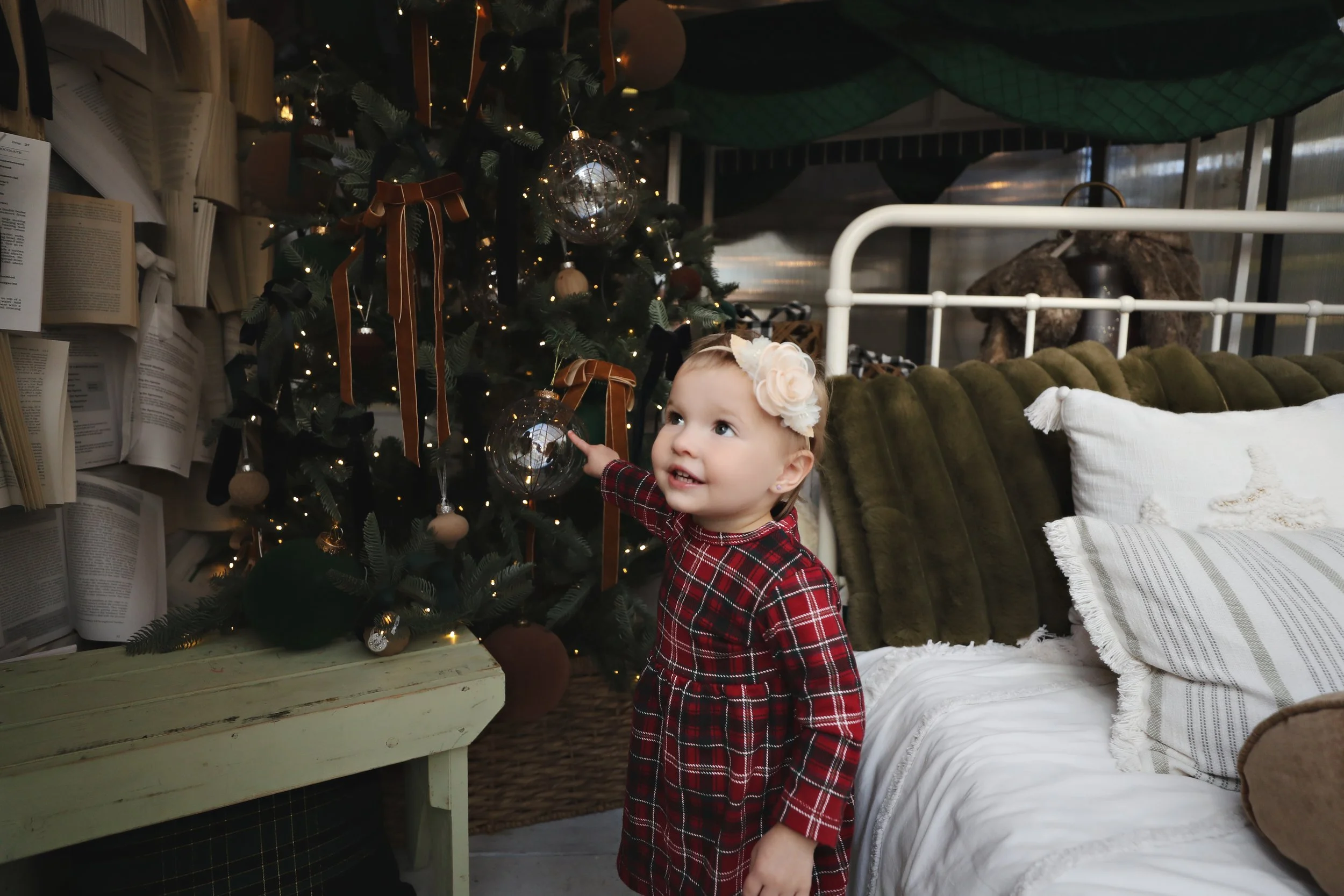 A young girl in a red plaid dress with a white flower headband decorating a Christmas tree with glass ornaments and ribbons inside a cozy living room.