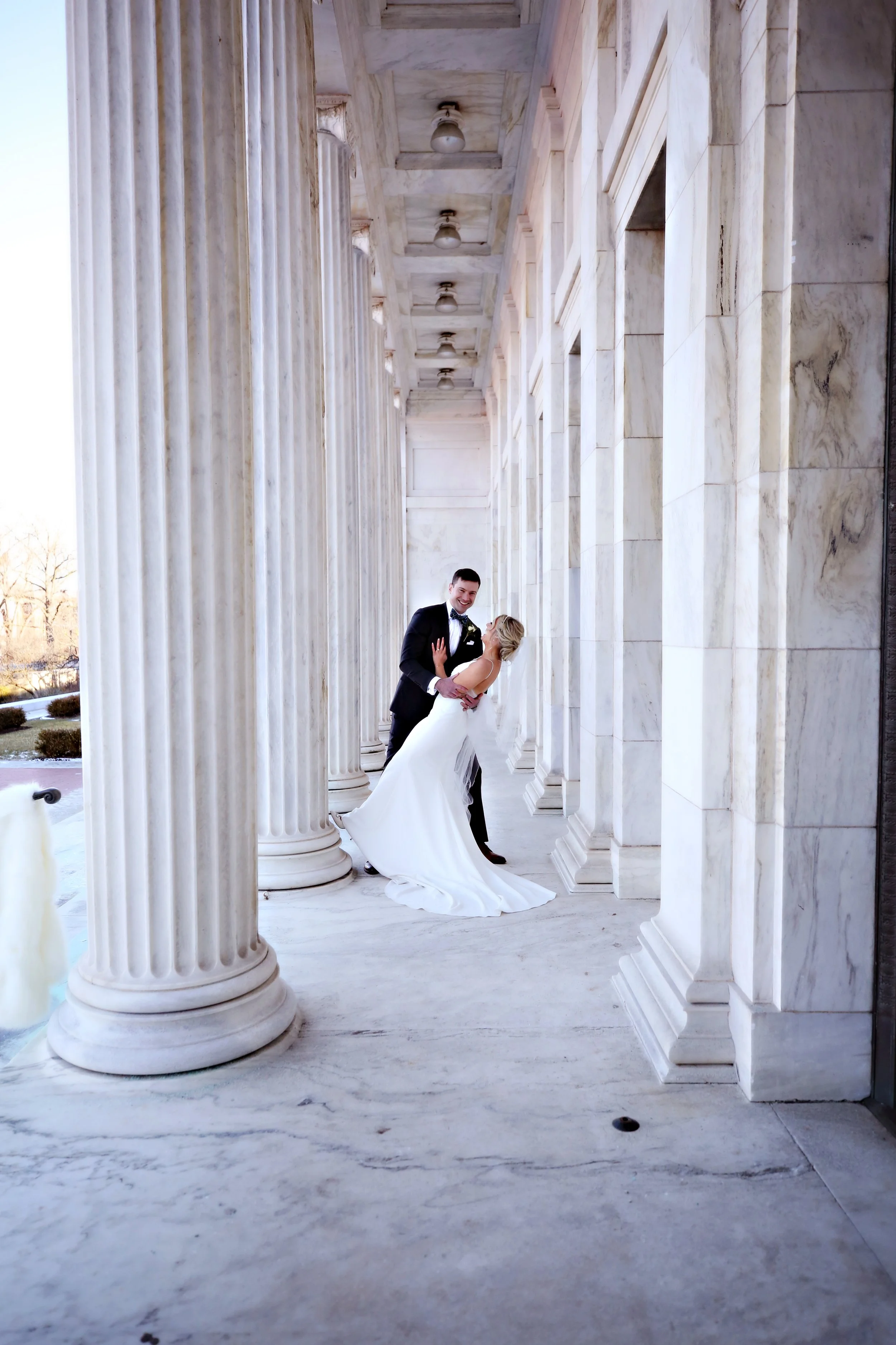 A newlywed couple in wedding attire posing and dancing in front of marble columns on a building porch.