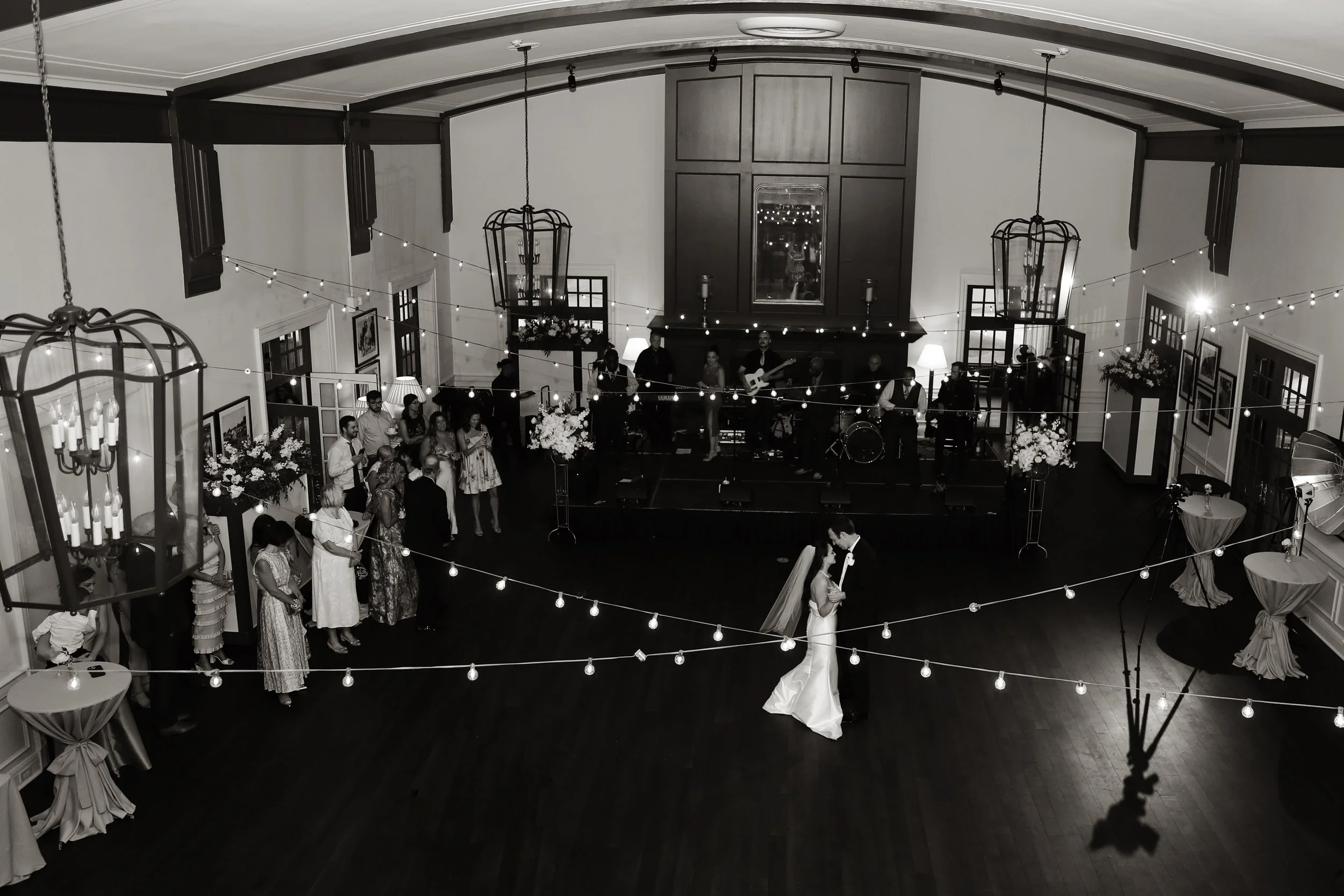 A black and white photo of a wedding reception with a bride and groom dancing in the center. Guests are gathered around, and a band plays music on a stage at the back of the room. Decorative string lights hang from the ceiling, creating a festive atm