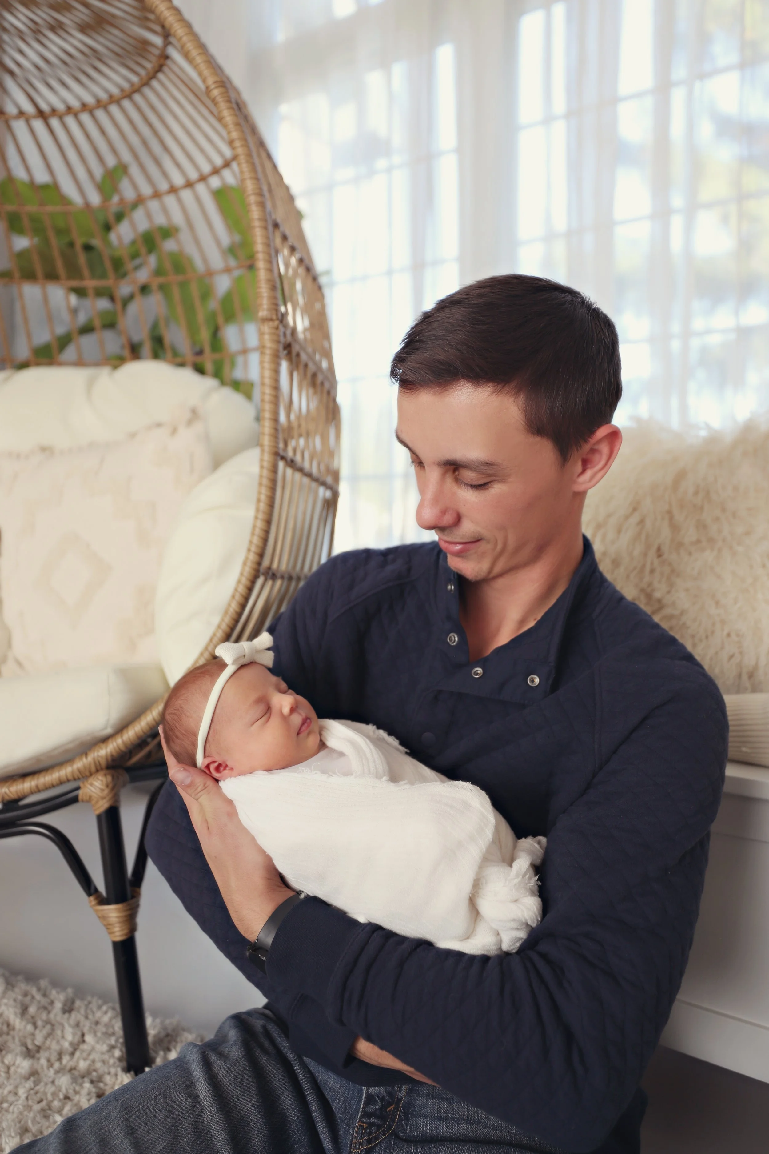 A man holding a sleeping baby in his arms inside a cozy room with bright natural light.