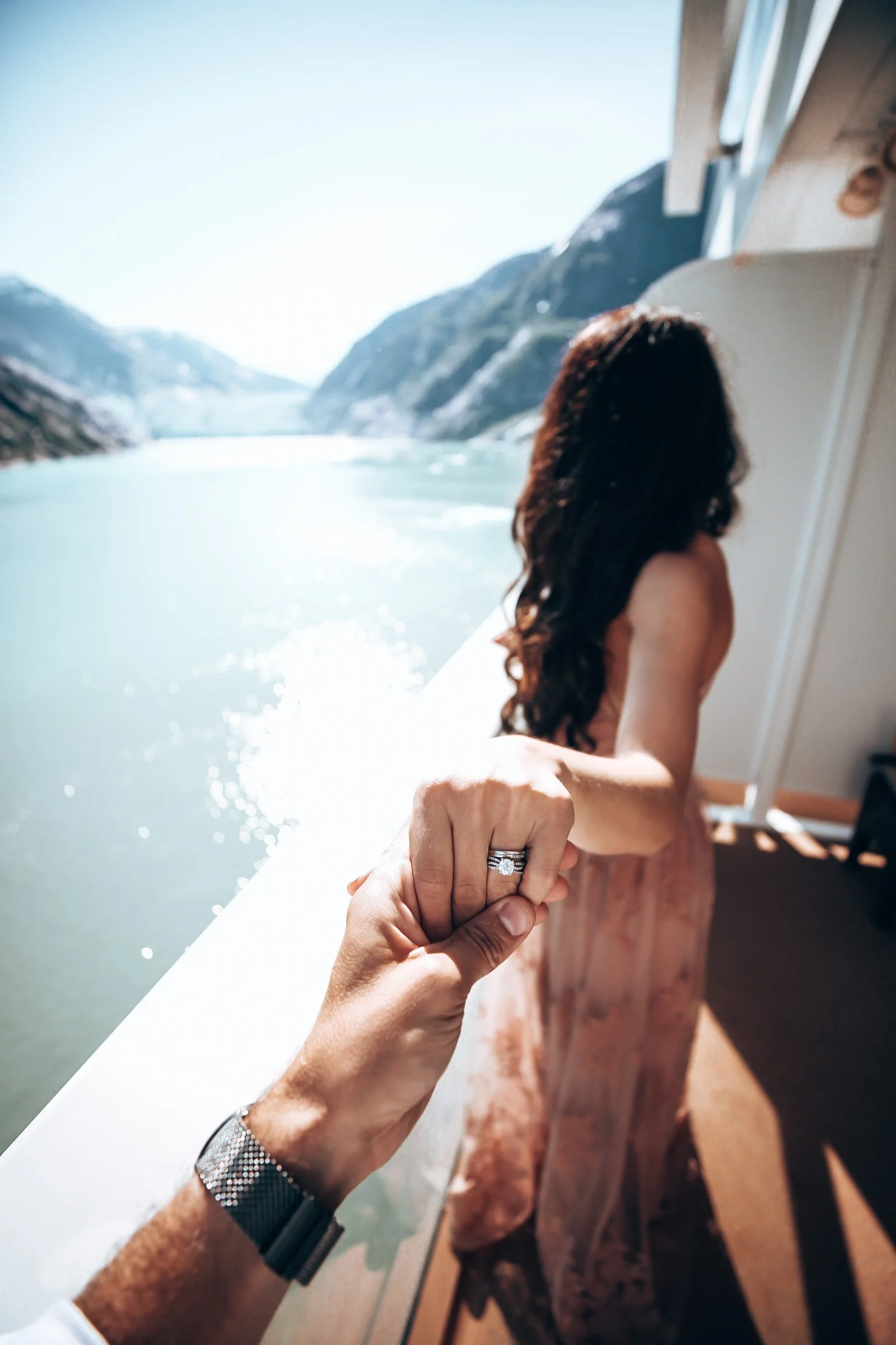 A couple holding hands on a boat with a large body of water and snow-covered mountains in the background, showing a close-up of a diamond engagement ring on the woman's finger.
