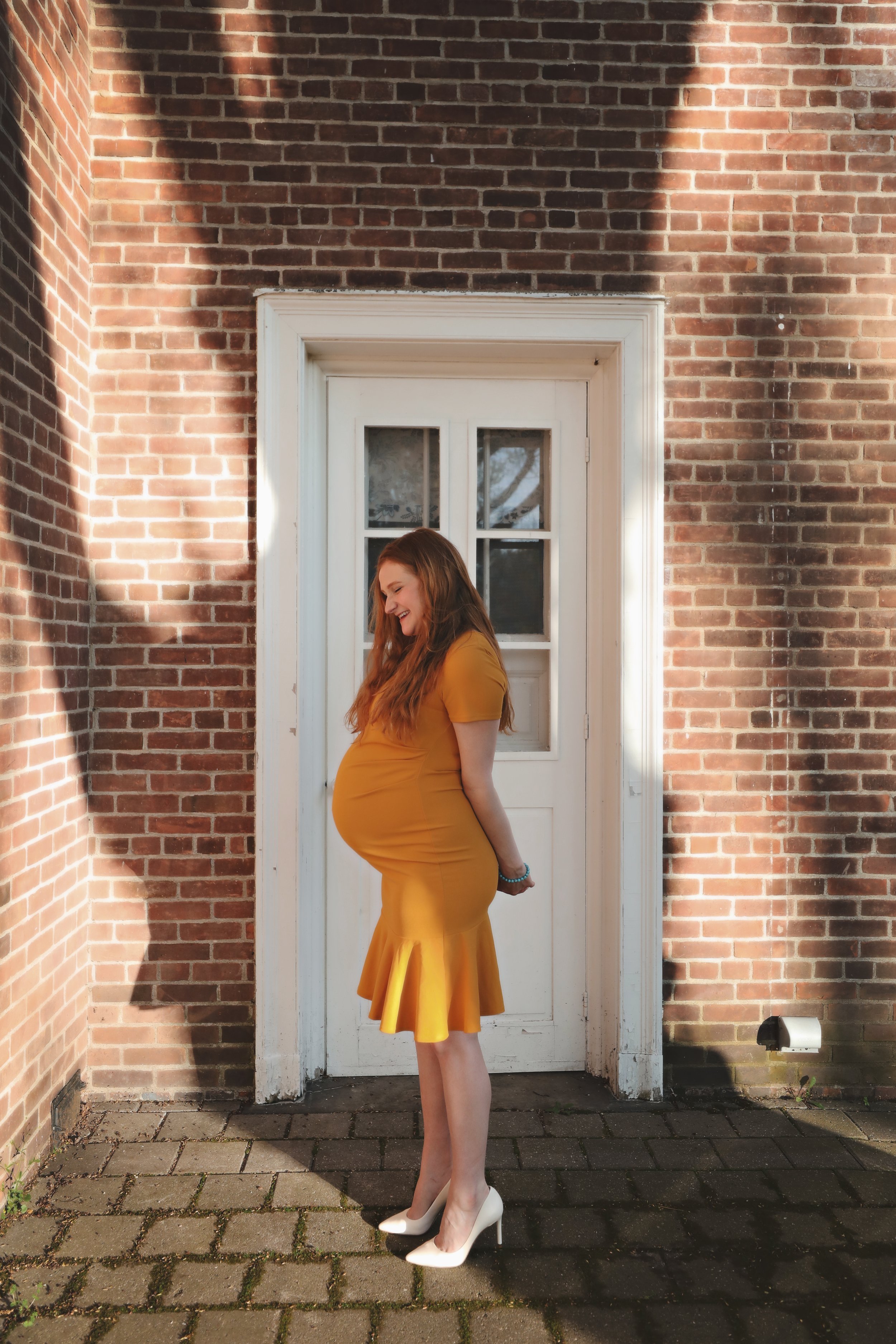 Pregnant woman in a yellow dress and white high heels standing outside near a brick wall and white door, smiling and looking down.