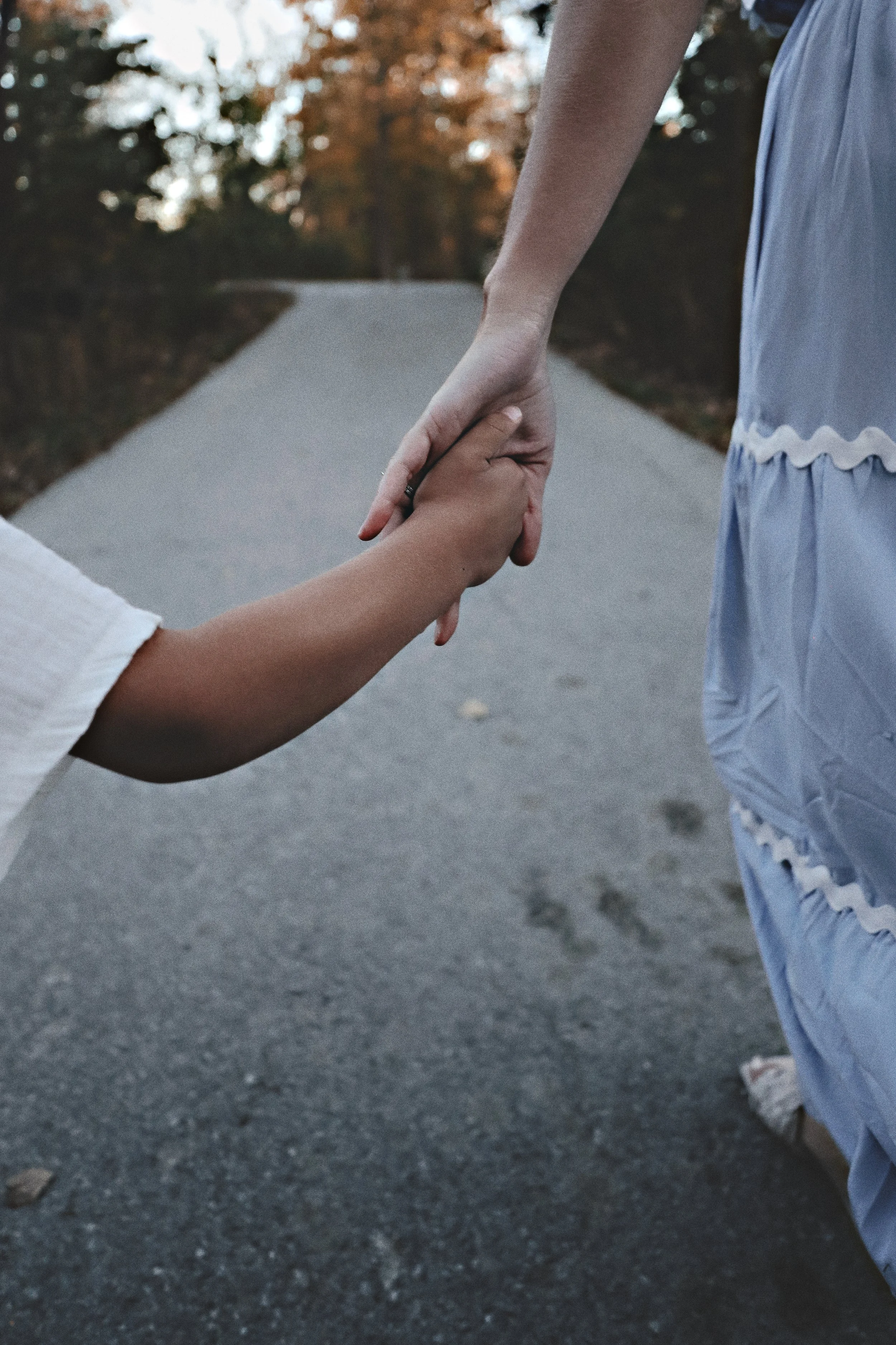 A person in a white dress holds the hand of another person in a blue dress on a gray pavement with trees in the background.