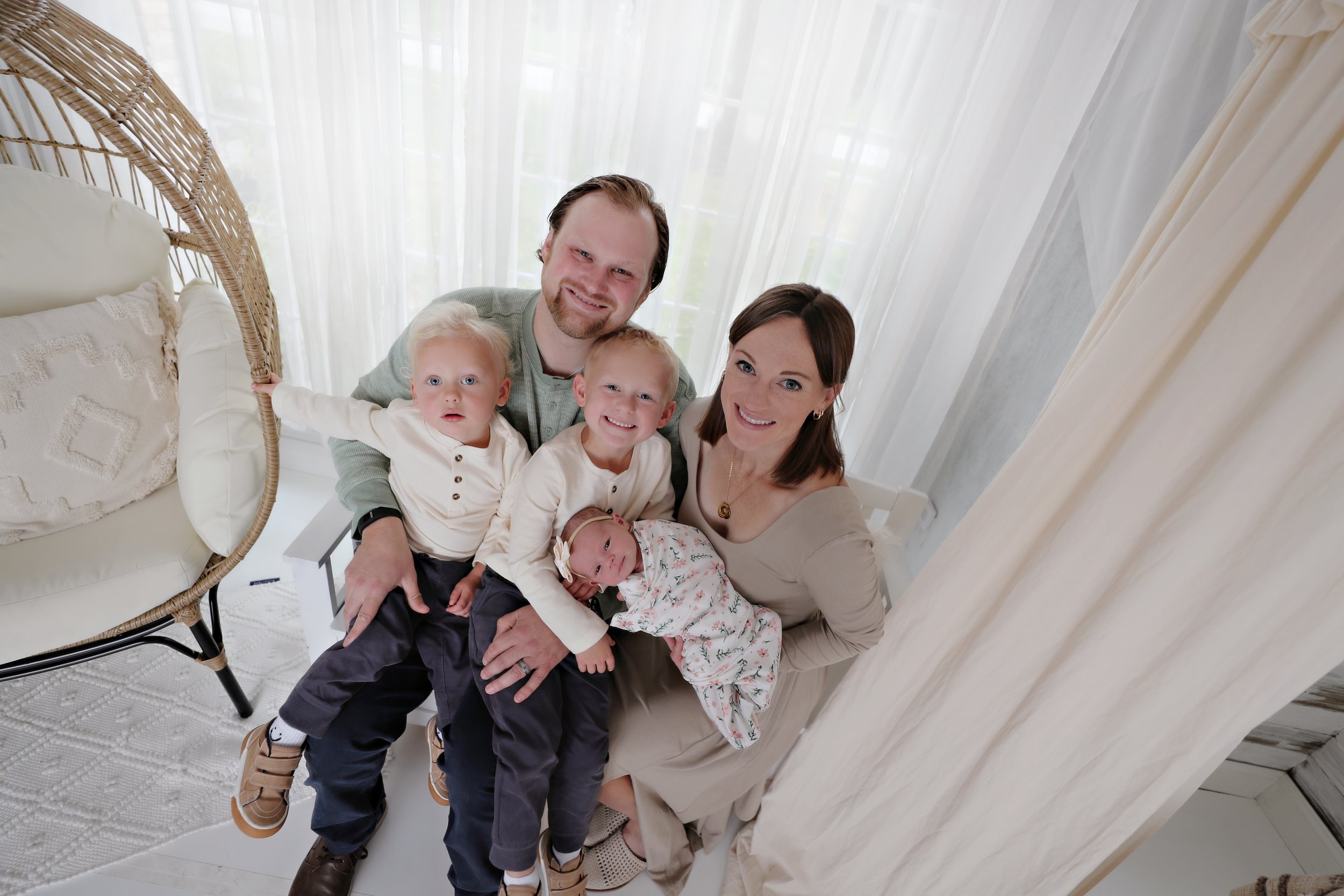 Family of five taking a selfie in a bright room. The family includes a man, woman, two young boys, and a newborn baby.