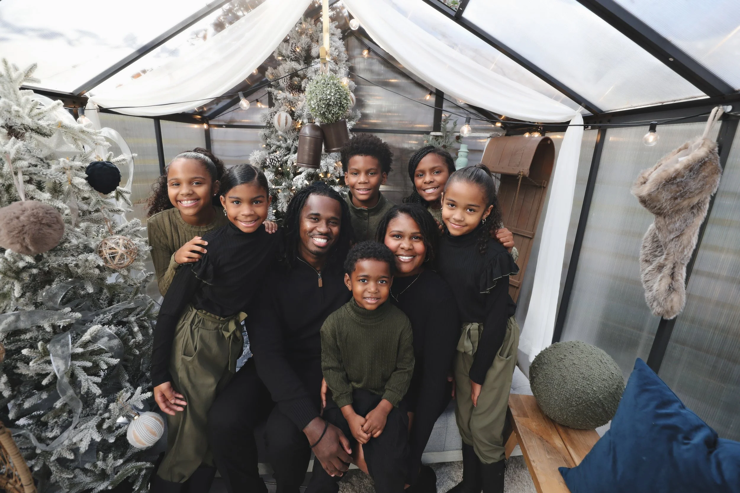 A family gathering of ten, including children and adults, smiling and posing inside a decorated greenhouse with Christmas decorations, a Christmas tree, and a stocking hanging on the wall.